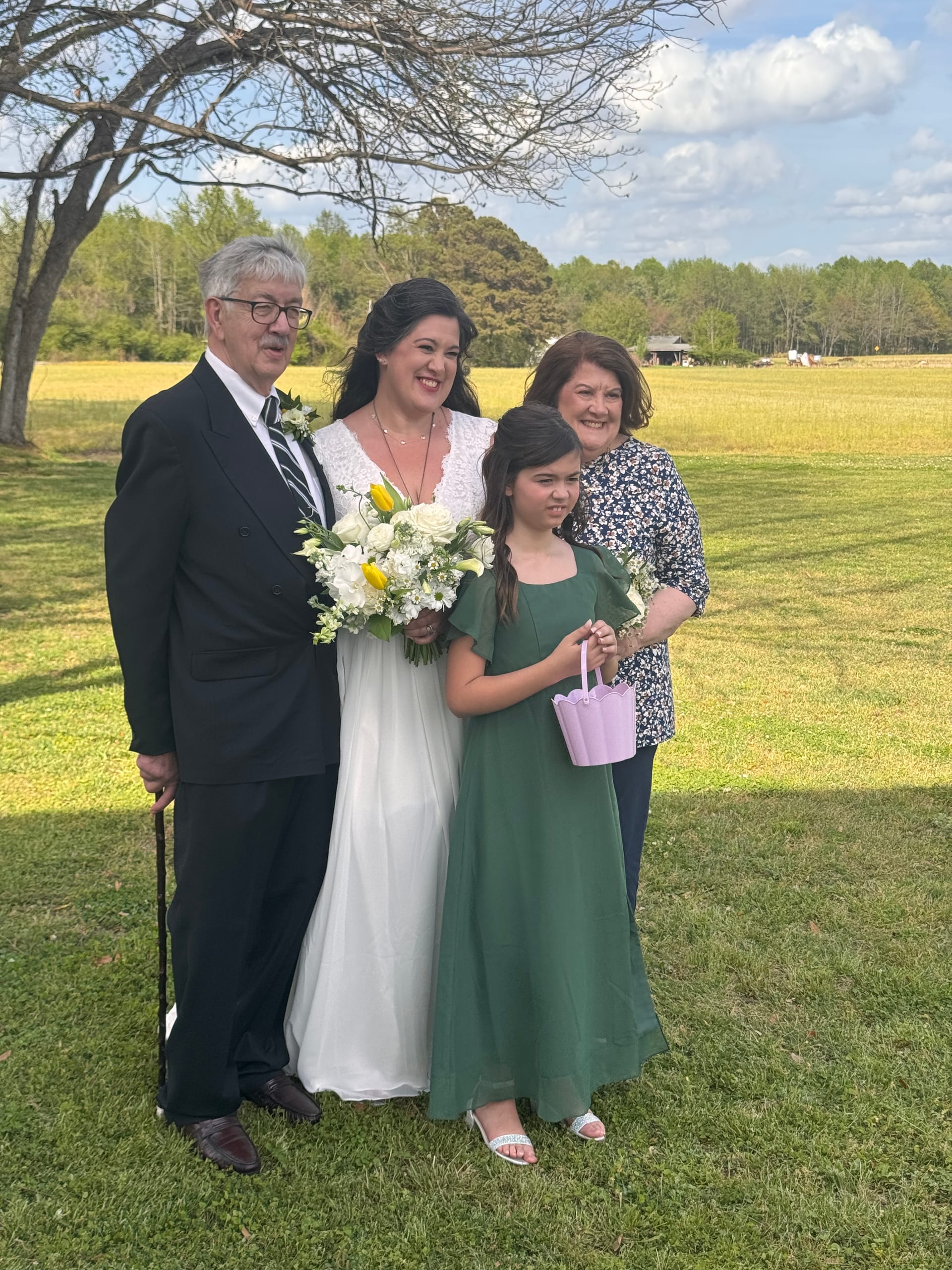 Bride in white gown holding a white and yellow bouquet poses with a young flower girl in green dress and two family members on the ceremony lawn with golden farmland fields in the background at Scarborough House.