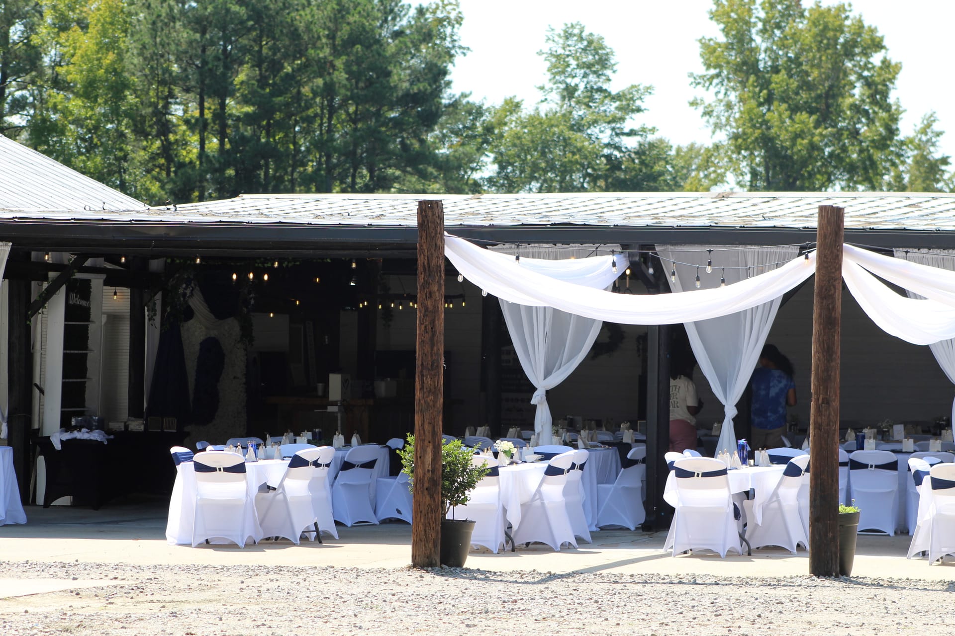 Elegant outdoor wedding reception setup at Scarborough House magnolia pavilion featuring white-draped wooden posts, round tables with white linens and navy chair sashes, and warm string lights.