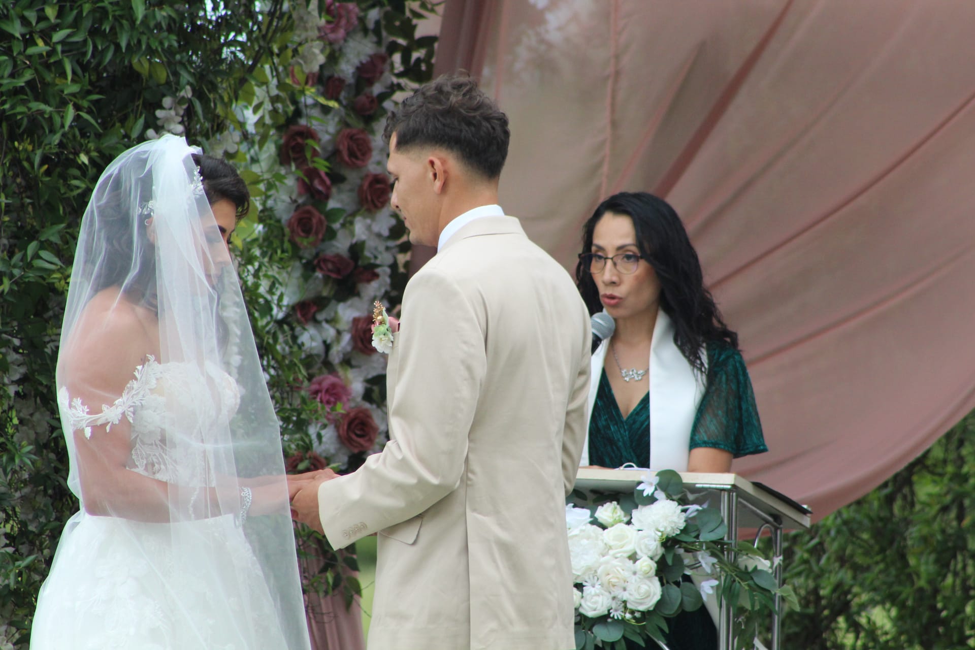 Bride and groom hold hands during their outdoor wedding ceremony at Scarborough House as the officiant speaks at a podium adorned with white roses against a lush floral arch backdrop.