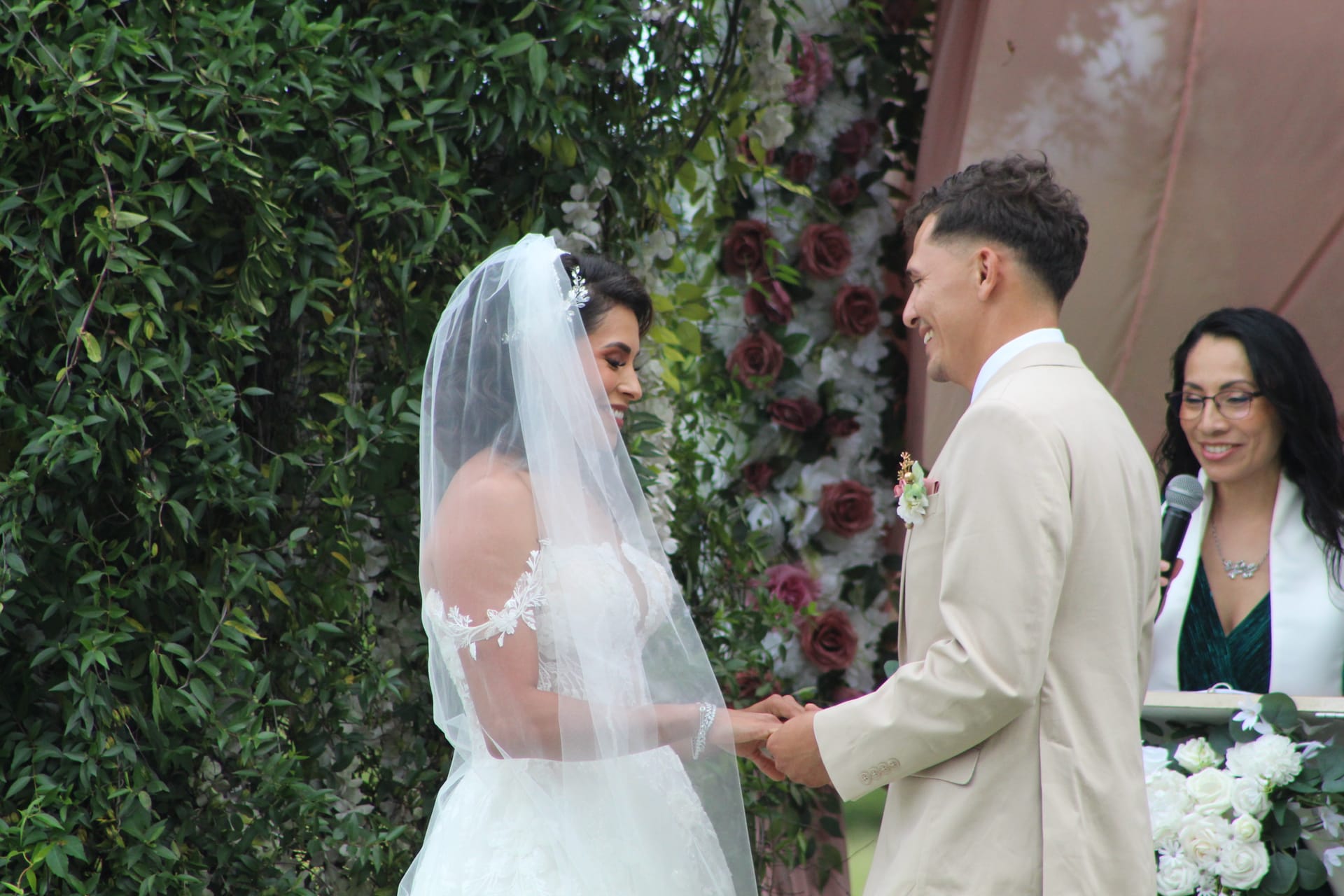 Bride in white lace gown and veil holds hands with smiling groom in tan suit during outdoor wedding ceremony before a lush floral arch backdrop.