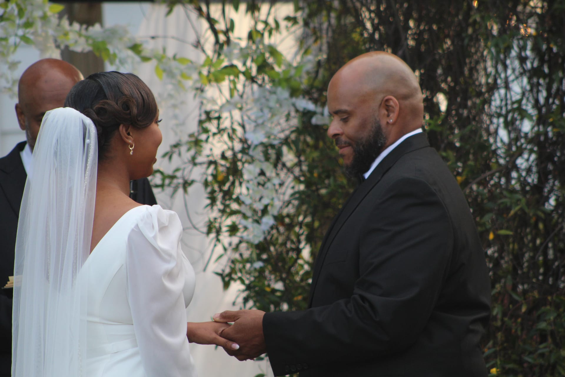 Bride in a white gown and veil holds hands with her groom in a black suit during an outdoor wedding ceremony at Scarborough House surrounded by lush greenery.