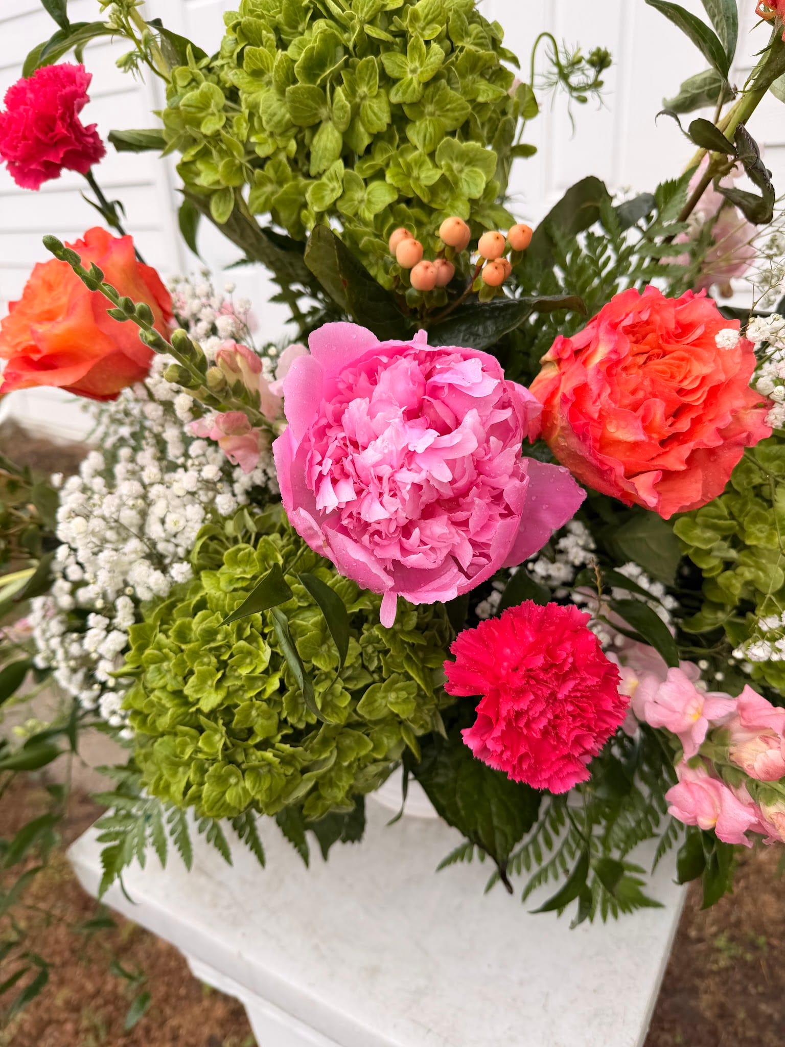 Lush wedding floral arrangement in a white urn featuring a pink peony, coral roses, red carnations, green hydrangeas, baby's breath, and hypericum berries at Scarborough House estate.