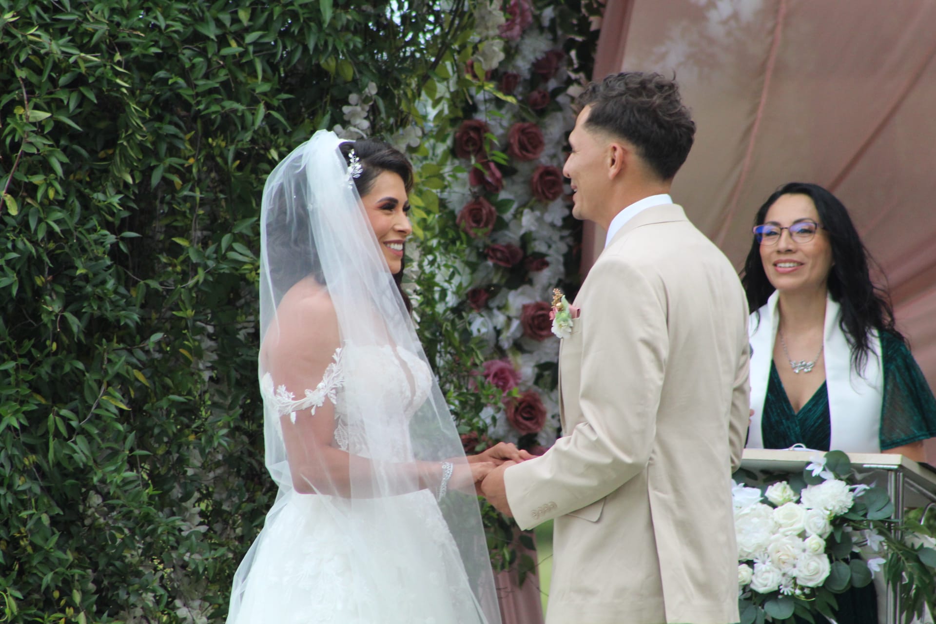 Bride in a lace gown and veil smiles joyfully at the groom in a tan suit as they hold hands during their outdoor wedding ceremony in front of a lush floral arch.
