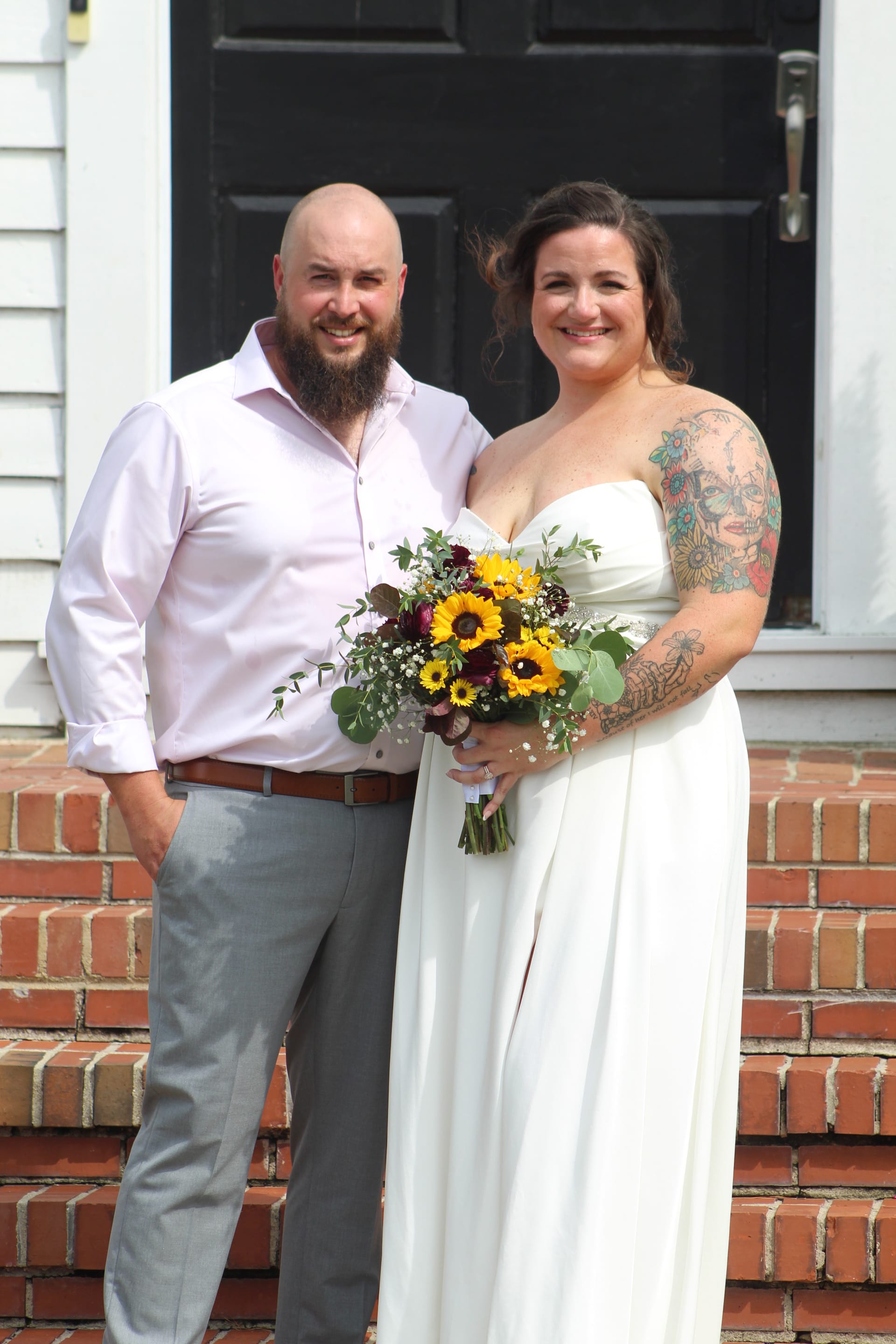 Smiling bride in a white strapless gown holding a sunflower bouquet stands with her bearded groom in a light pink shirt on the brick front steps of the historic 1821 Scarborough House estate.