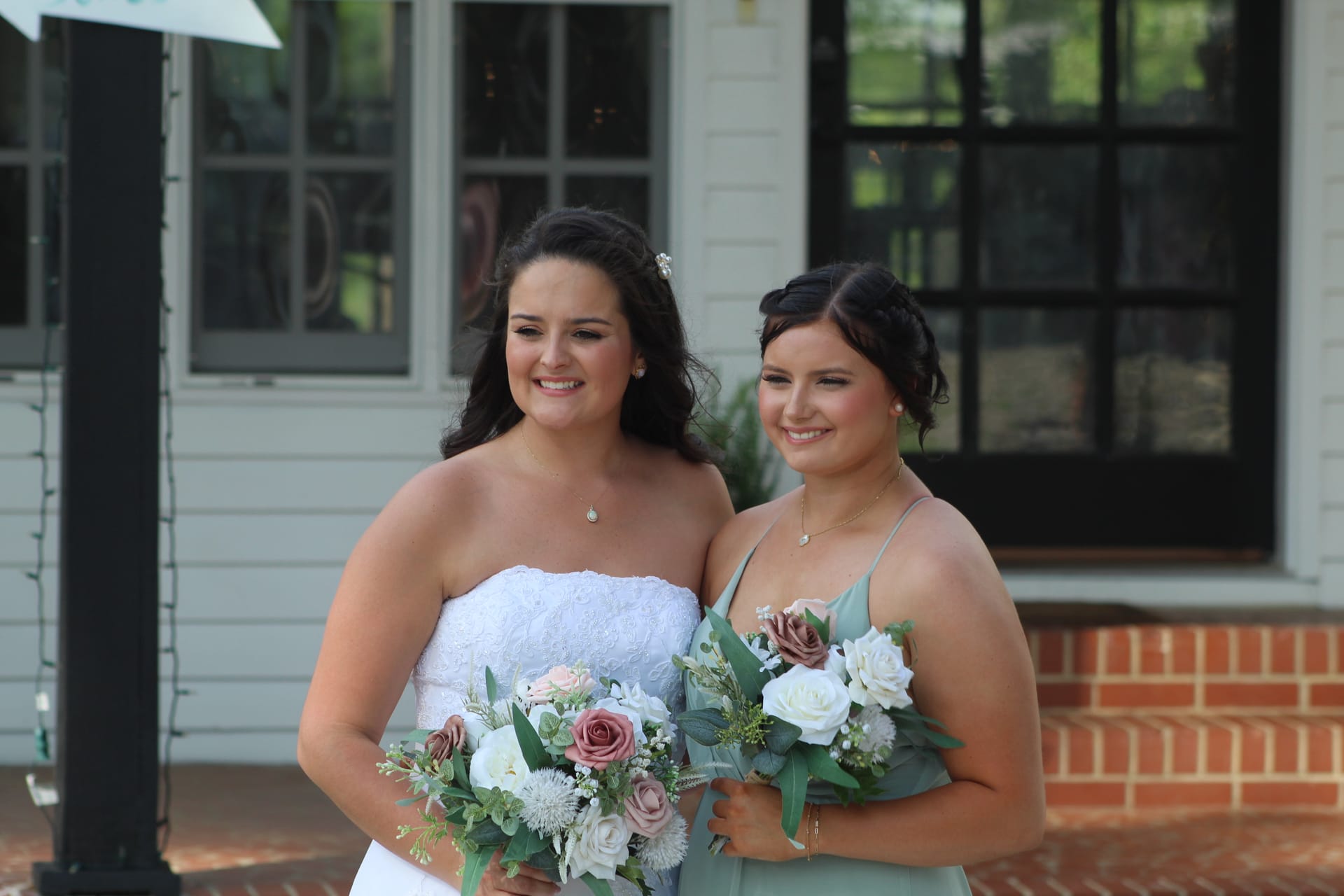 Bride in a strapless white lace gown and bridesmaid in sage green holding floral bouquets of dusty rose and white roses in front of the historic Scarborough House manor.