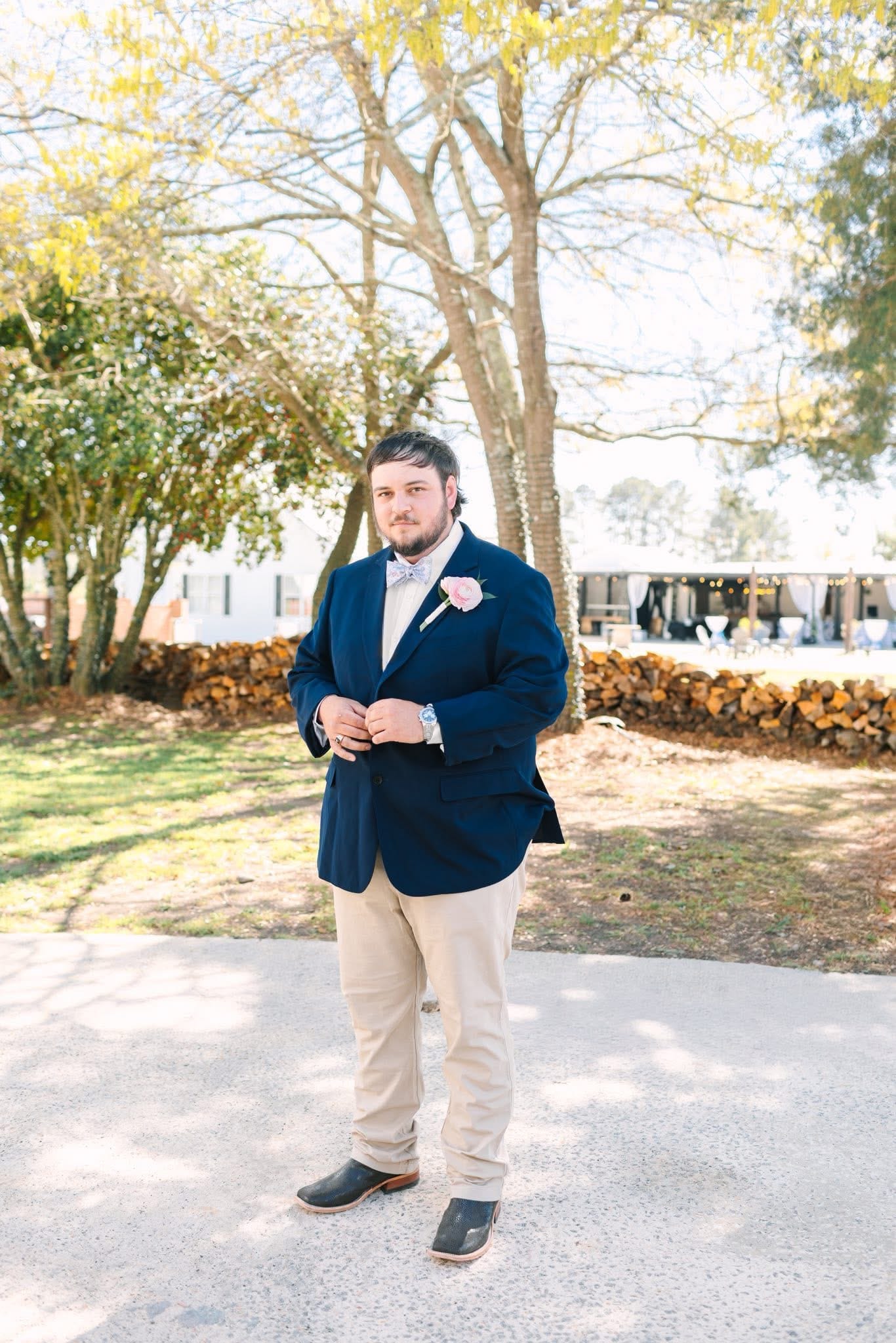 Groom in a navy blazer with a pink boutonniere and bow tie stands confidently on the grounds of Scarborough House wedding venue surrounded by trees and stacked firewood.