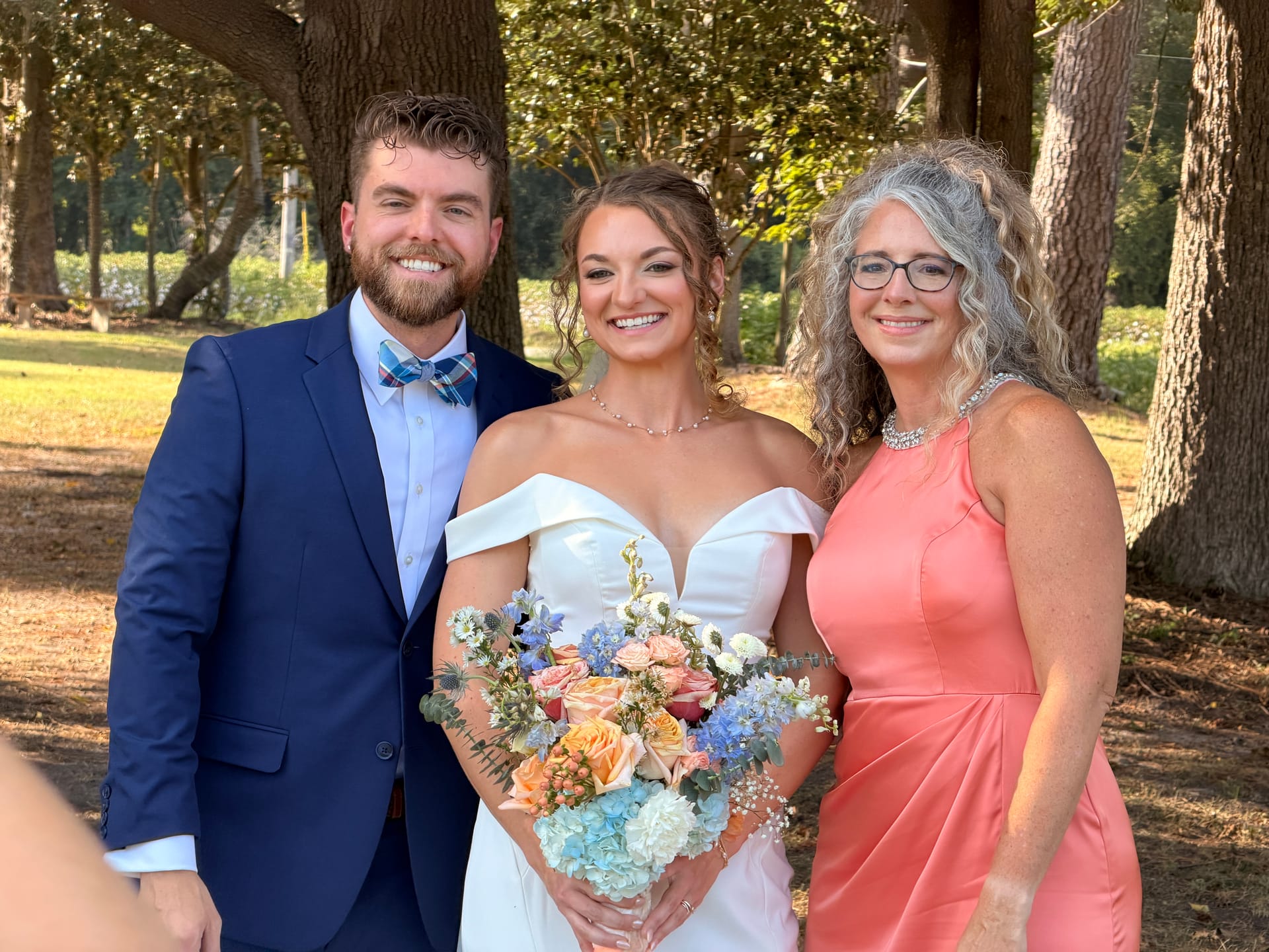 Bride in off-shoulder white gown holding a pastel bouquet poses with groom in navy suit and bow tie and an older woman in coral dress beneath oak trees at Scarborough House.