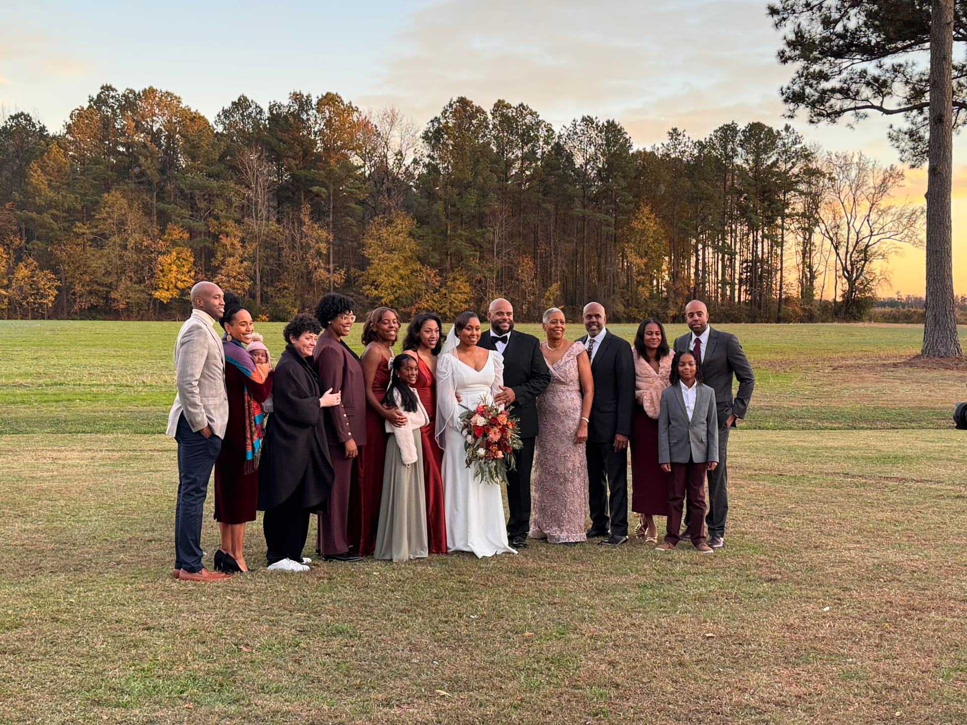 Bride and groom surrounded by smiling family members in burgundy and formal attire during a golden-hour sunset portrait on the open farmland fields at Scarborough House.