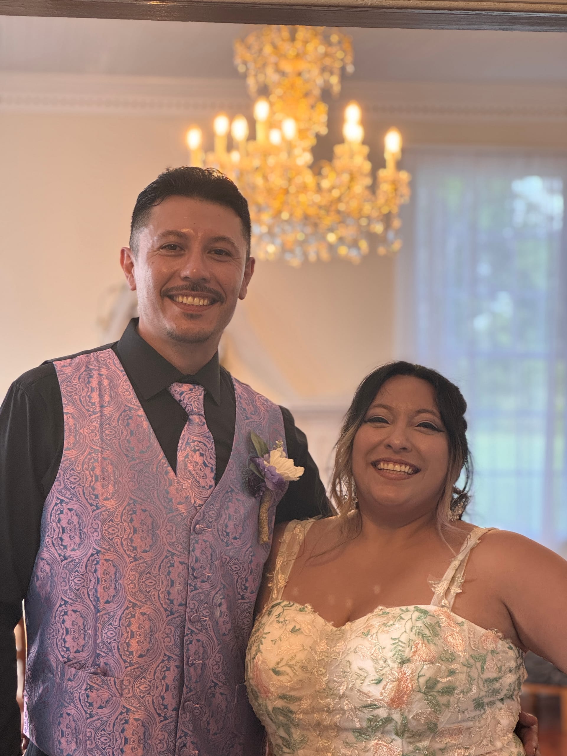 Smiling bride in a floral embroidered gown and groom in a purple paisley vest and boutonniere pose together beneath a glowing chandelier inside the grand hall at Scarborough House.