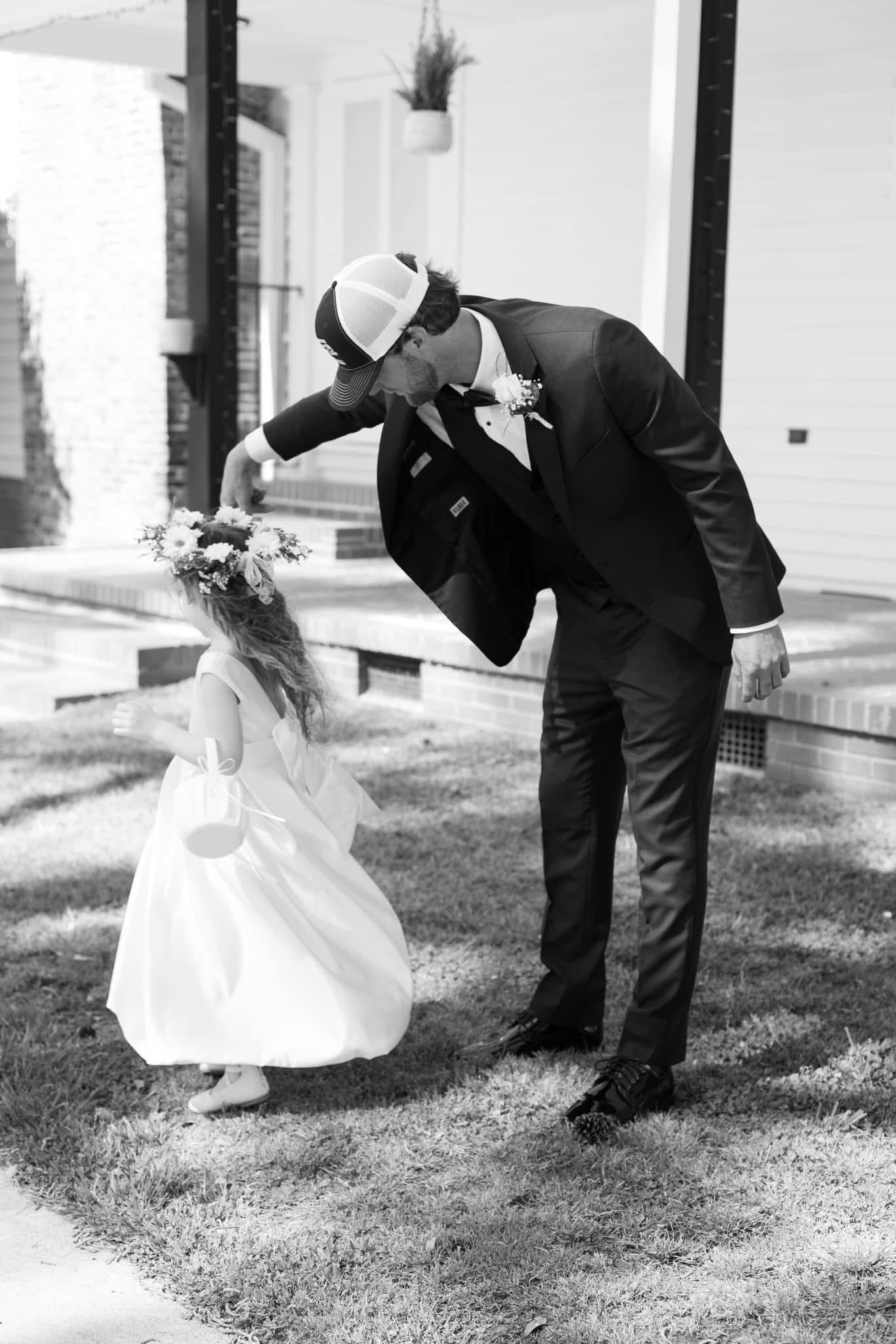 Groom in a dark suit and trucker cap bows playfully toward a young flower girl in a white dress and floral crown on the ceremony lawn at Scarborough House.