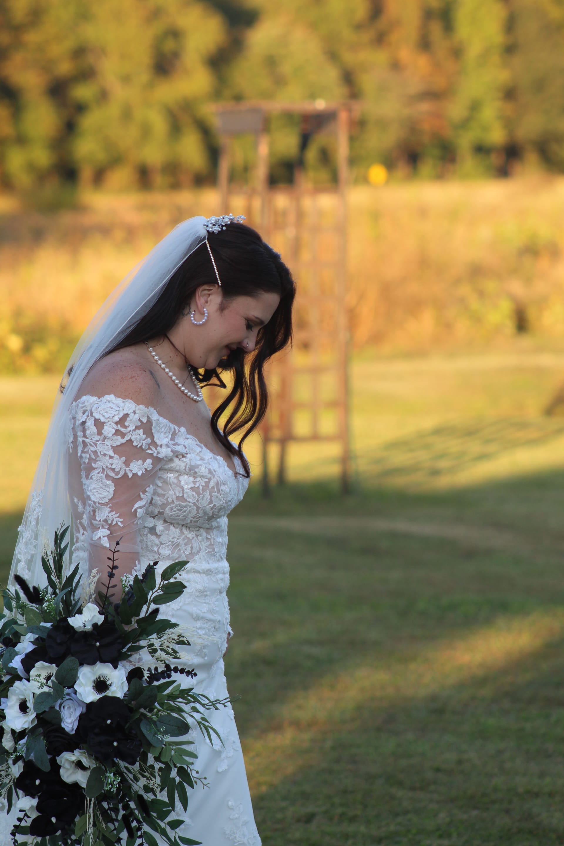 A bride in a long-sleeve lace gown and pearl necklace gazes downward holding a dramatic cascading bouquet of white anemones and dark foliage in warm golden-hour light on the ceremony lawn.