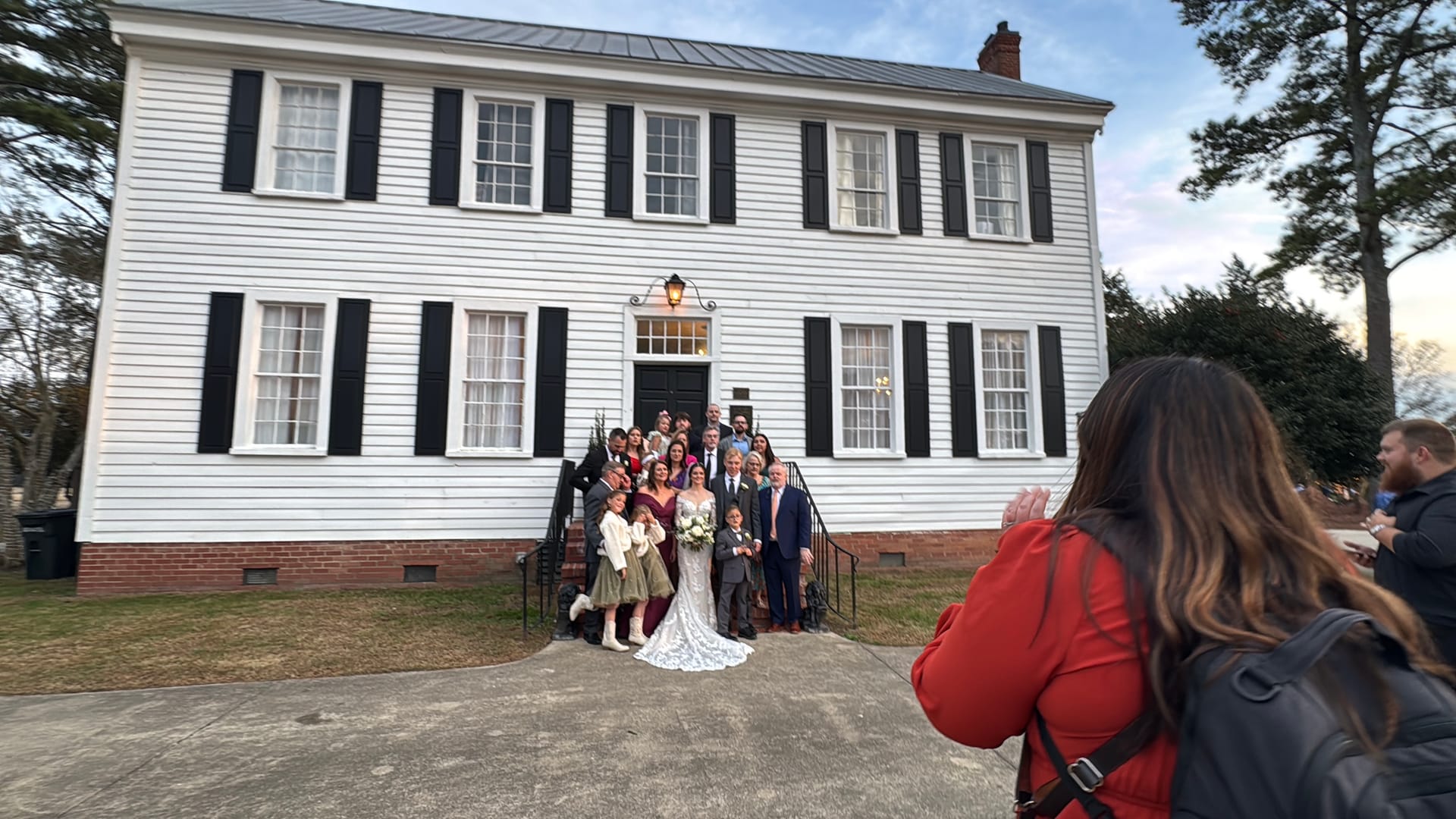 Wedding party and family gathered on the front steps of the 1821 Scarborough House white colonial manor at dusk, with the bride in a lace gown at center.