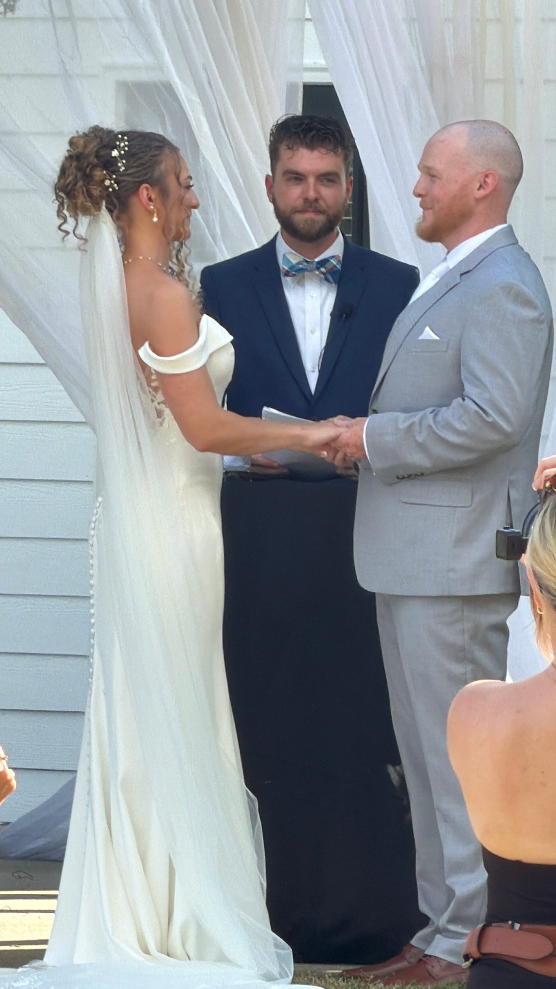 Bride and groom hold hands during their outdoor vow exchange before a white-draped backdrop at Scarborough House historic estate, officiated by a bearded man in a navy suit with a plaid bow tie.