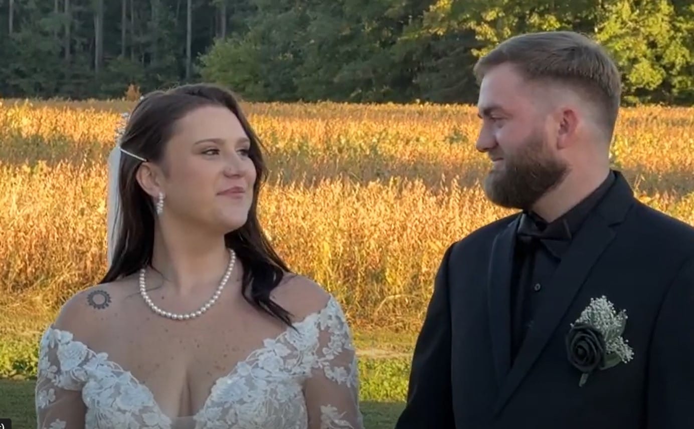 Bride in lace gown and groom in black tuxedo share a tender glance during golden-hour portraits in front of sunlit farmland fields at Scarborough House.