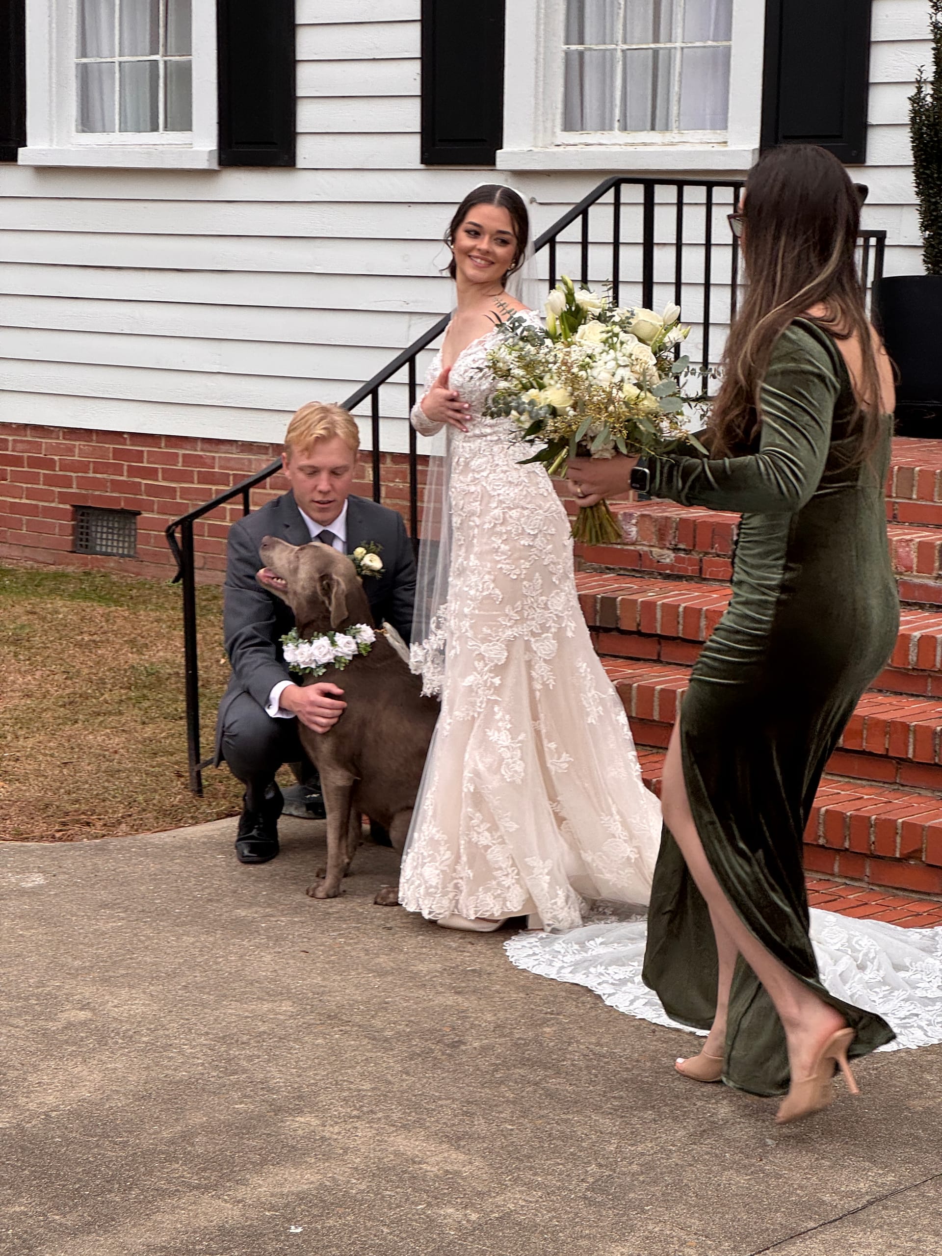 A radiant bride in a lace gown holds a white floral bouquet on the front steps of the historic Scarborough House estate while a groomsman crouches beside a dog wearing a floral collar and a bridesmaid in green velvet approaches.