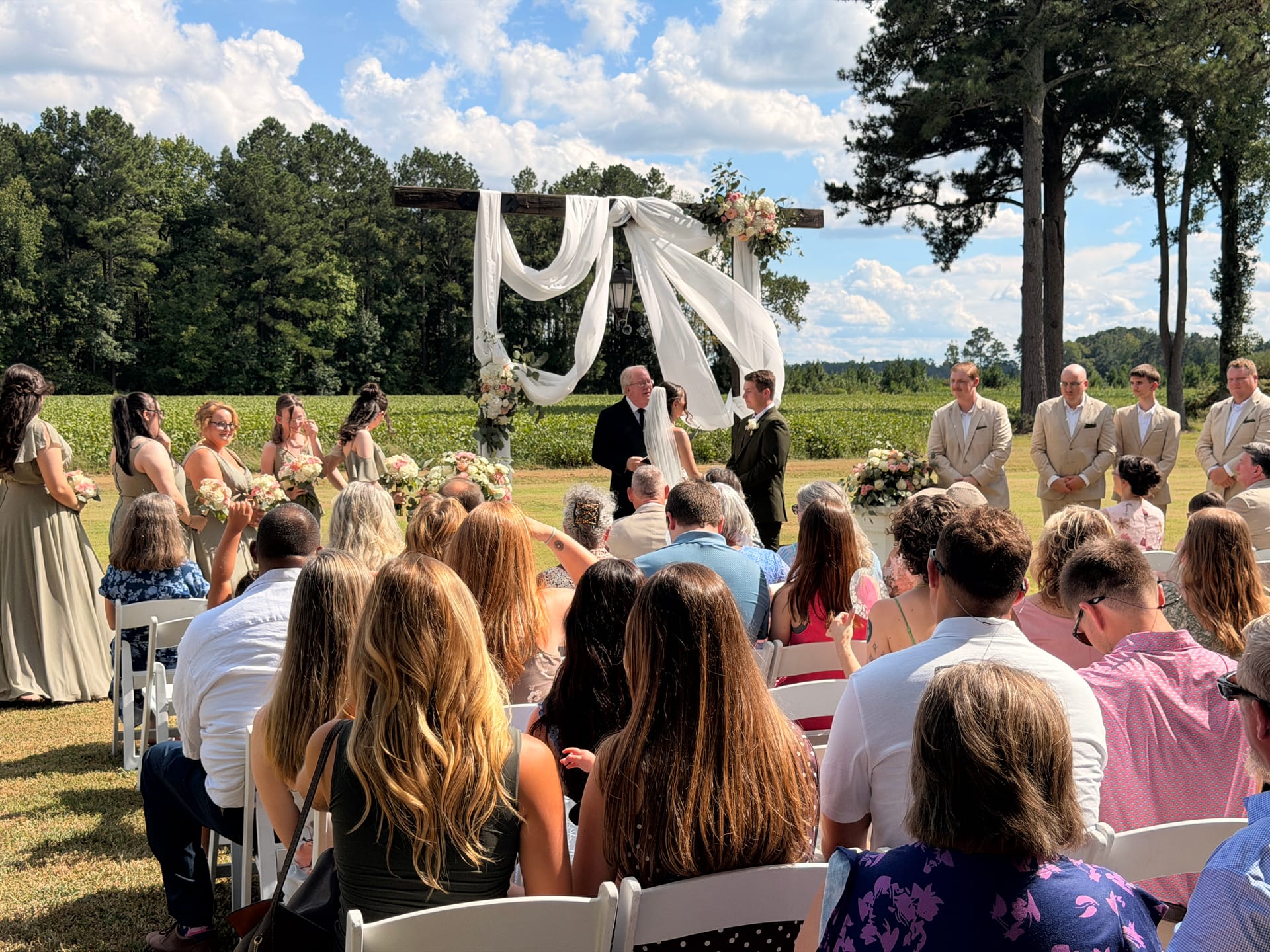 Guests seated in white chairs watch a couple exchange vows beneath a draped floral arch on the ceremony lawn at Scarborough House, with bridesmaids, groomsmen, and open farmland fields visible in the background.