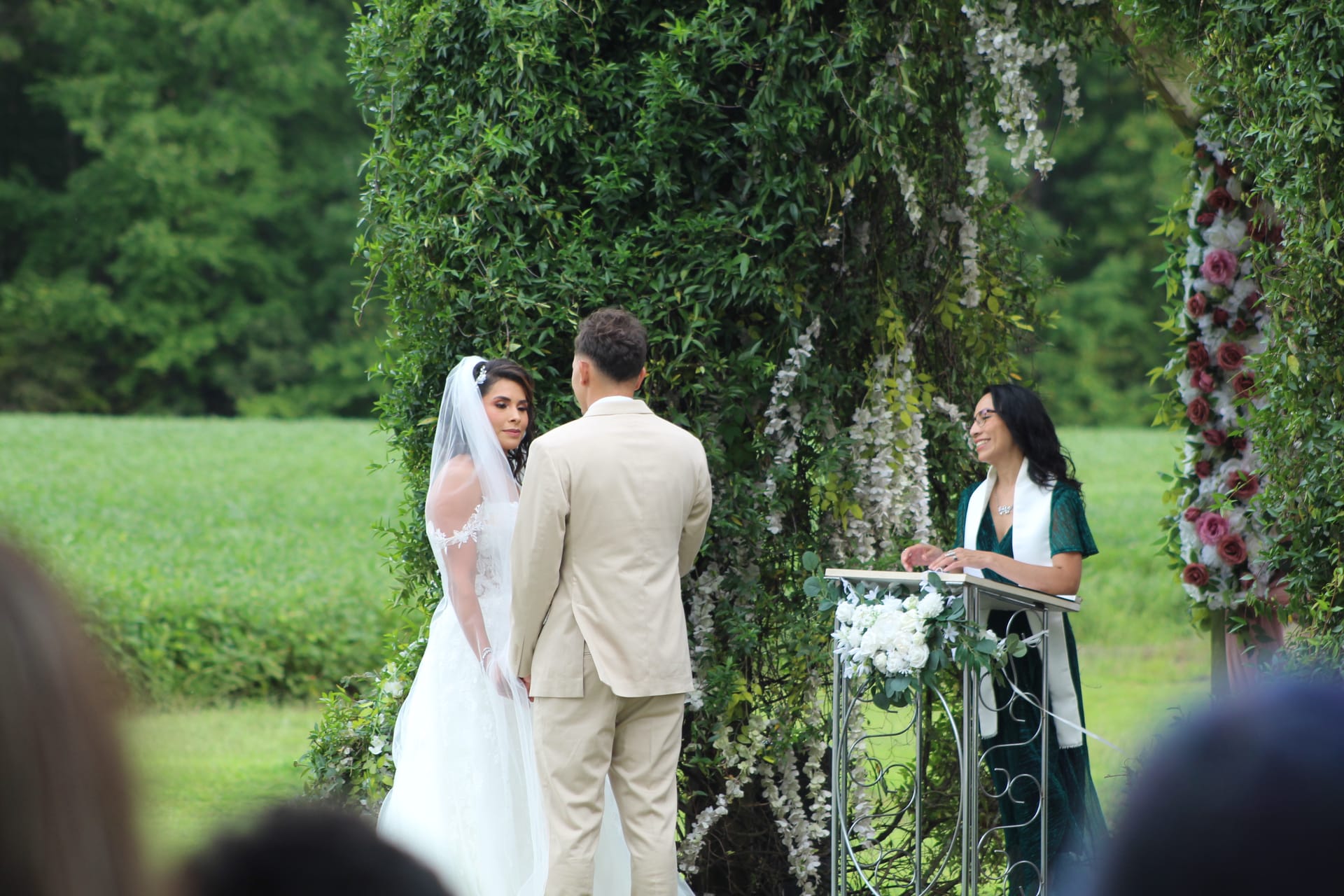 Bride in white gown and veil exchanges vows with groom beneath a lush floral and greenery arch as the officiant smiles at a podium on the ceremony lawn at Scarborough House.