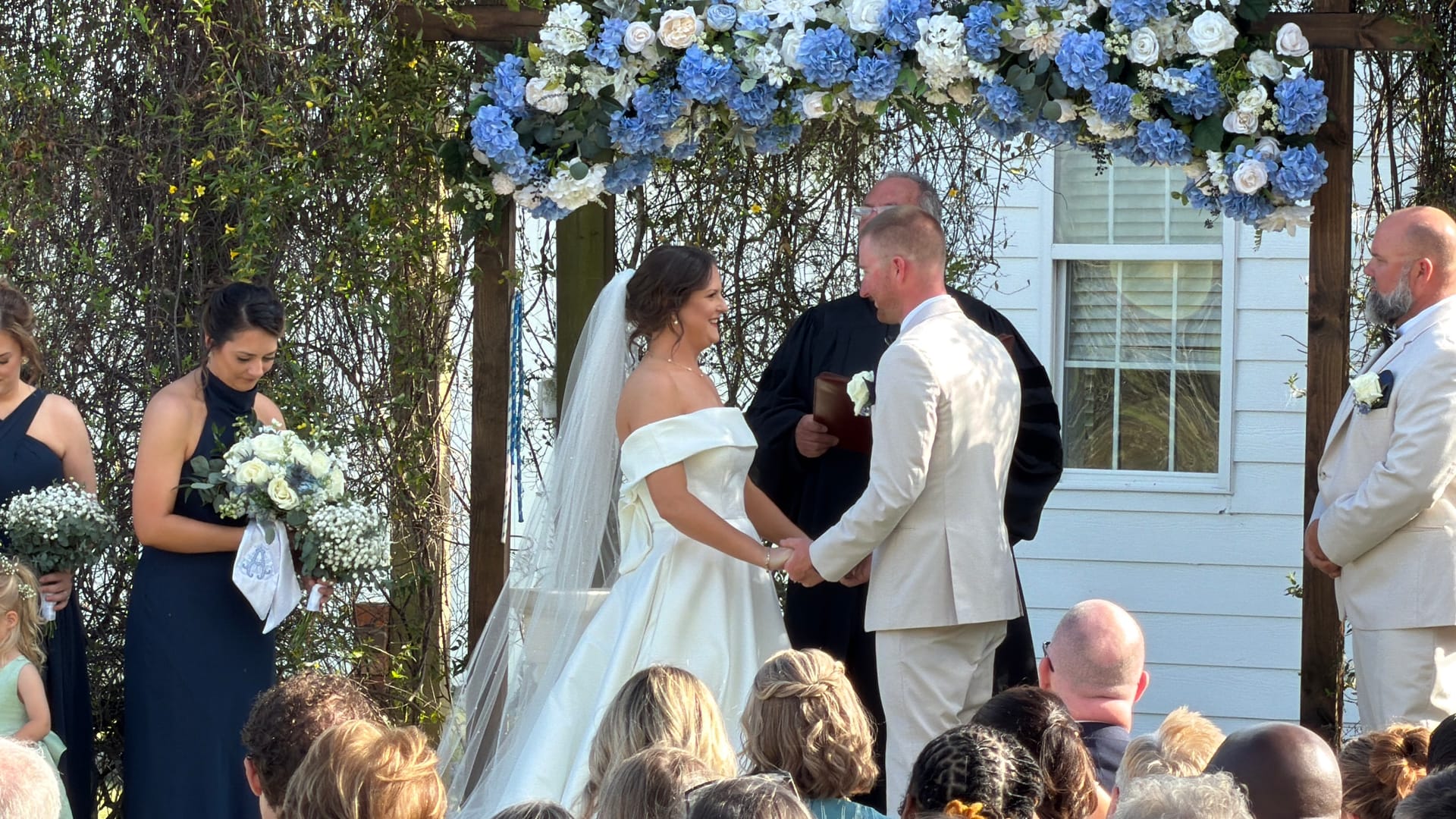 Bride in an off-shoulder white gown smiles at her groom in a cream suit as they exchange vows beneath a blue and white floral arch at Scarborough House wedding venue.