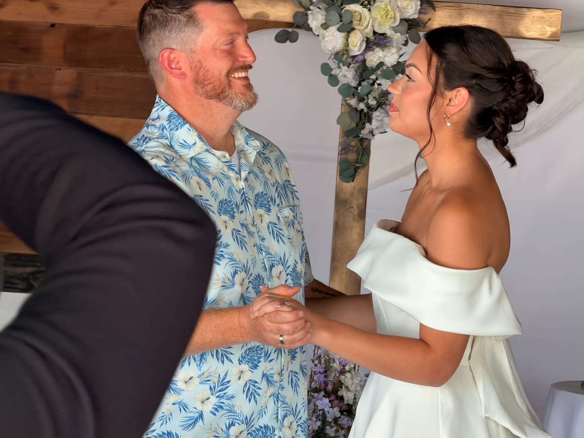 A laughing groom in a floral shirt holds hands with his smiling bride in an off-shoulder white gown beneath a wooden arch decorated with white roses and eucalyptus.