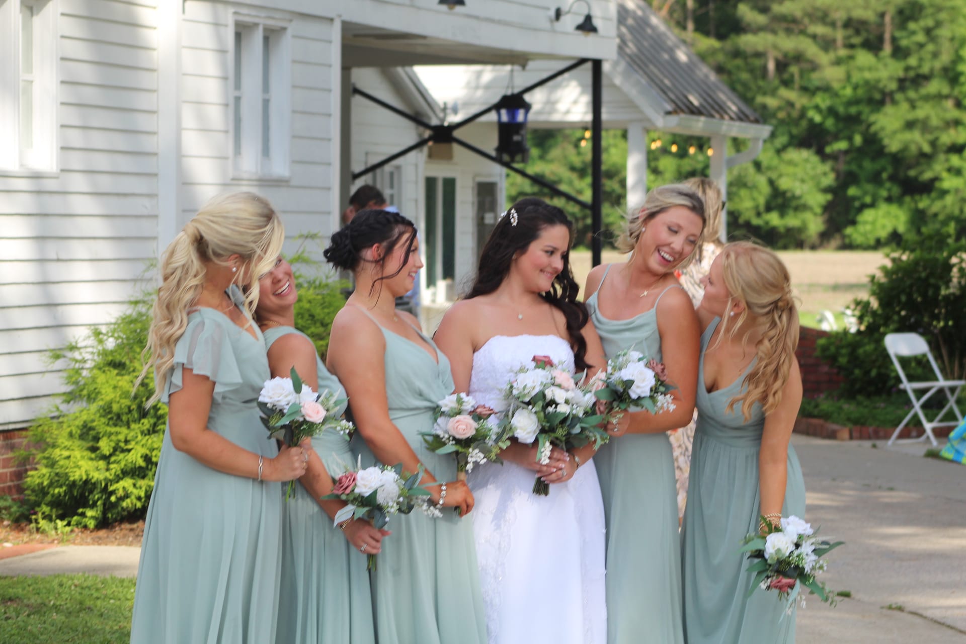 Bride in white strapless gown laughing with four bridesmaids in sage green dresses holding floral bouquets outside the historic Scarborough House manor.