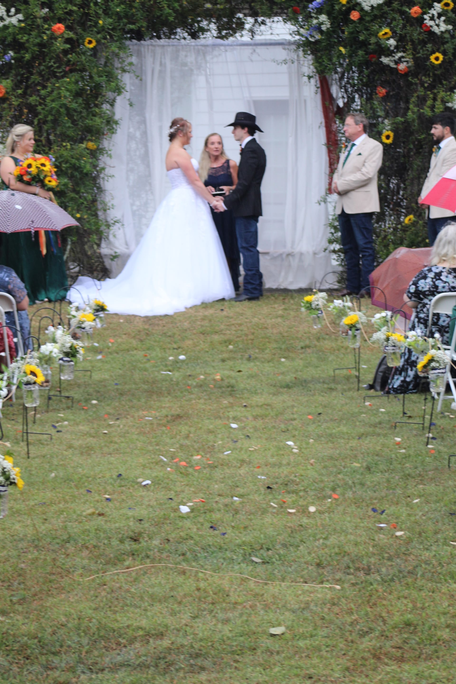 Bride in a white ball gown and groom in a black cowboy hat exchange vows beneath a floral-draped arch on the ceremony lawn at Scarborough House, flanked by sunflower-adorned aisle décor.