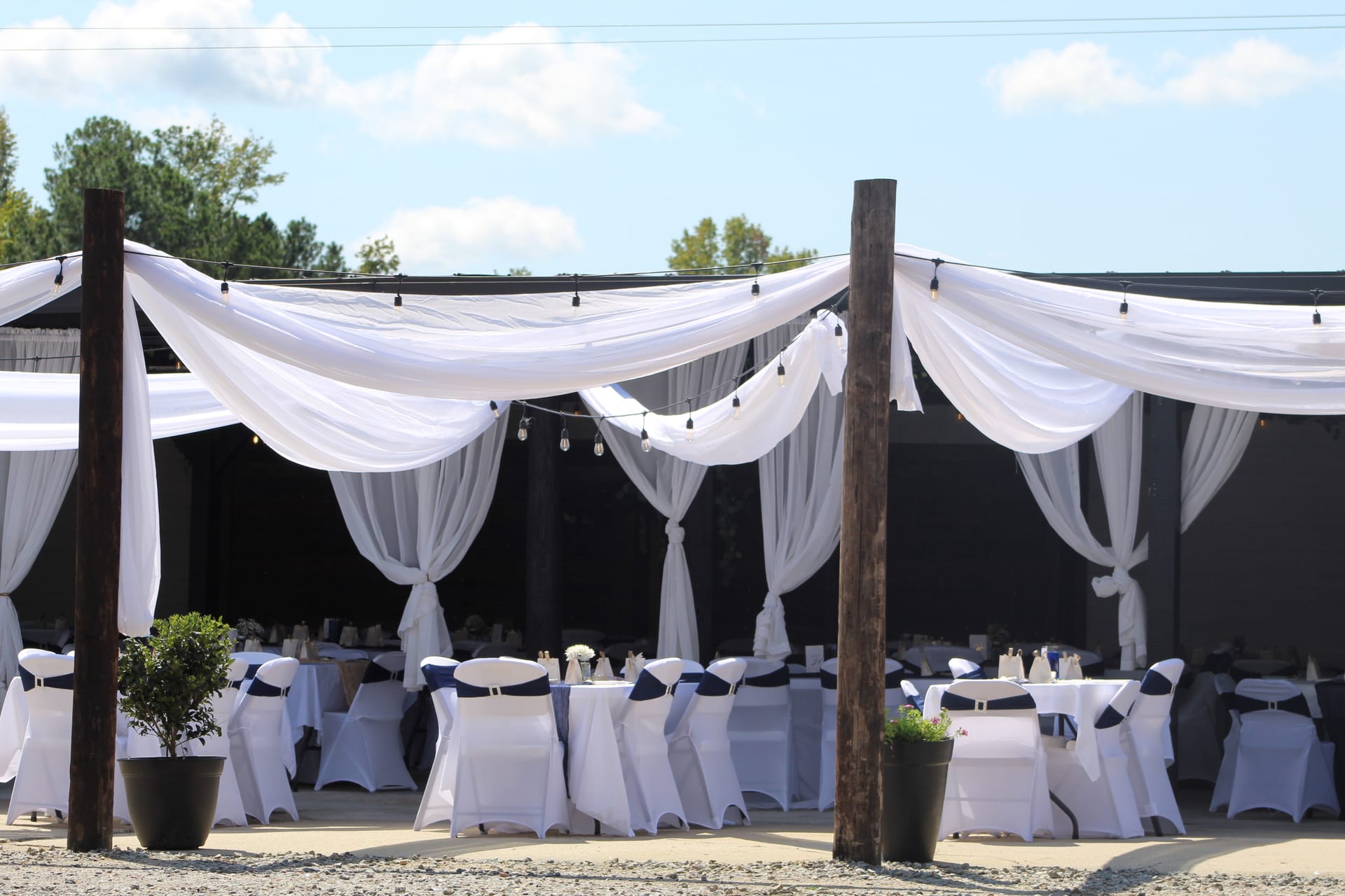 Elegant outdoor wedding reception at Scarborough House featuring sweeping white fabric draping, Edison string lights, and white chair-covered seats with navy sashes set beneath a rustic wooden pergola structure.