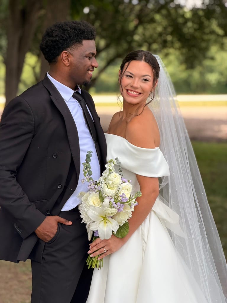 Bride in an off-shoulder white gown holding a white and lavender bouquet smiles at the camera while the groom in a black suit looks admiringly at her beneath towering oak trees at Scarborough House estate.