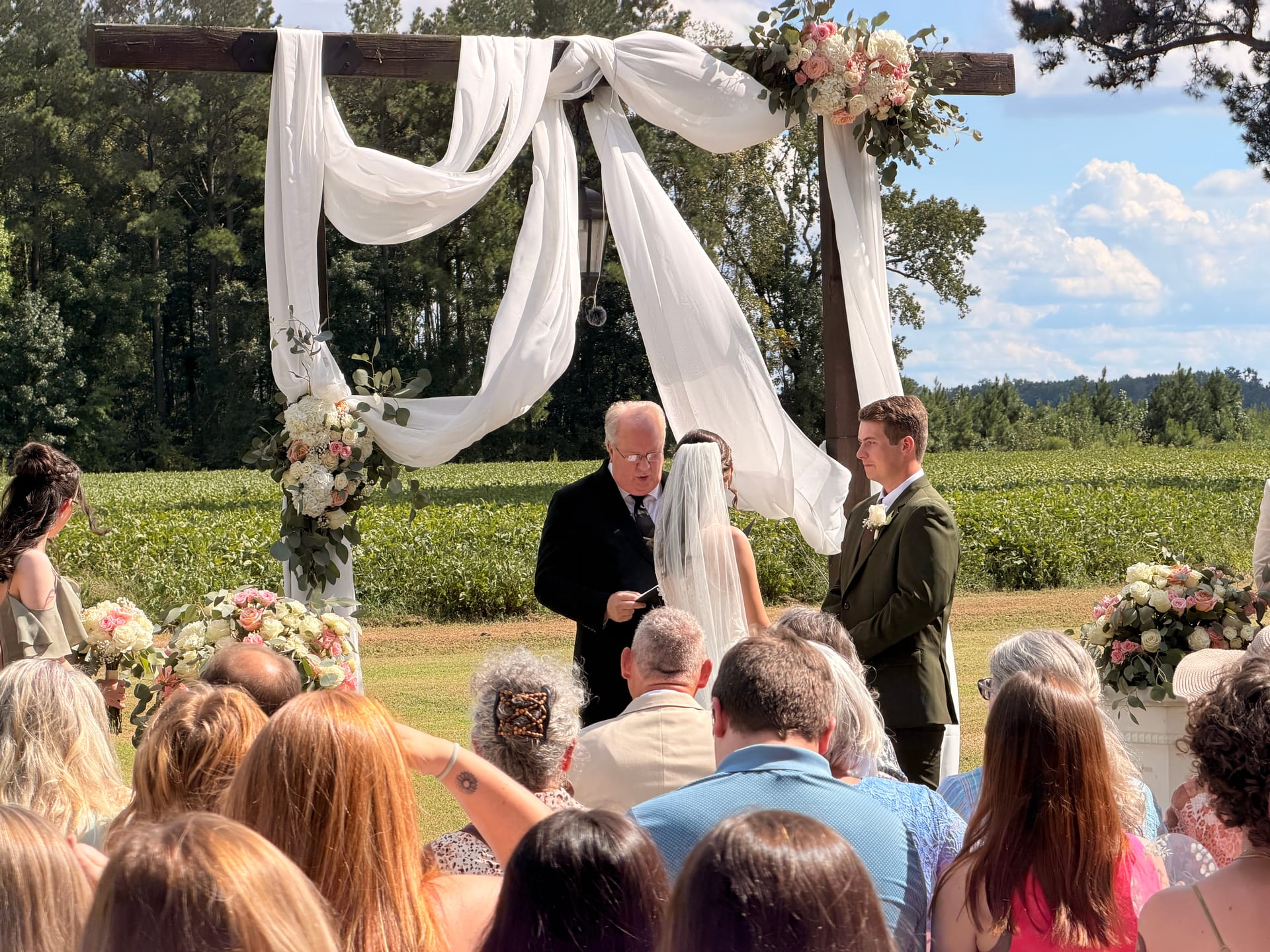 A groom in a dark green suit stands at a rustic wooden arch draped in white fabric and pink floral arrangements during an outdoor wedding ceremony on the Scarborough House ceremony lawn, with lush farmland fields visible in the background.