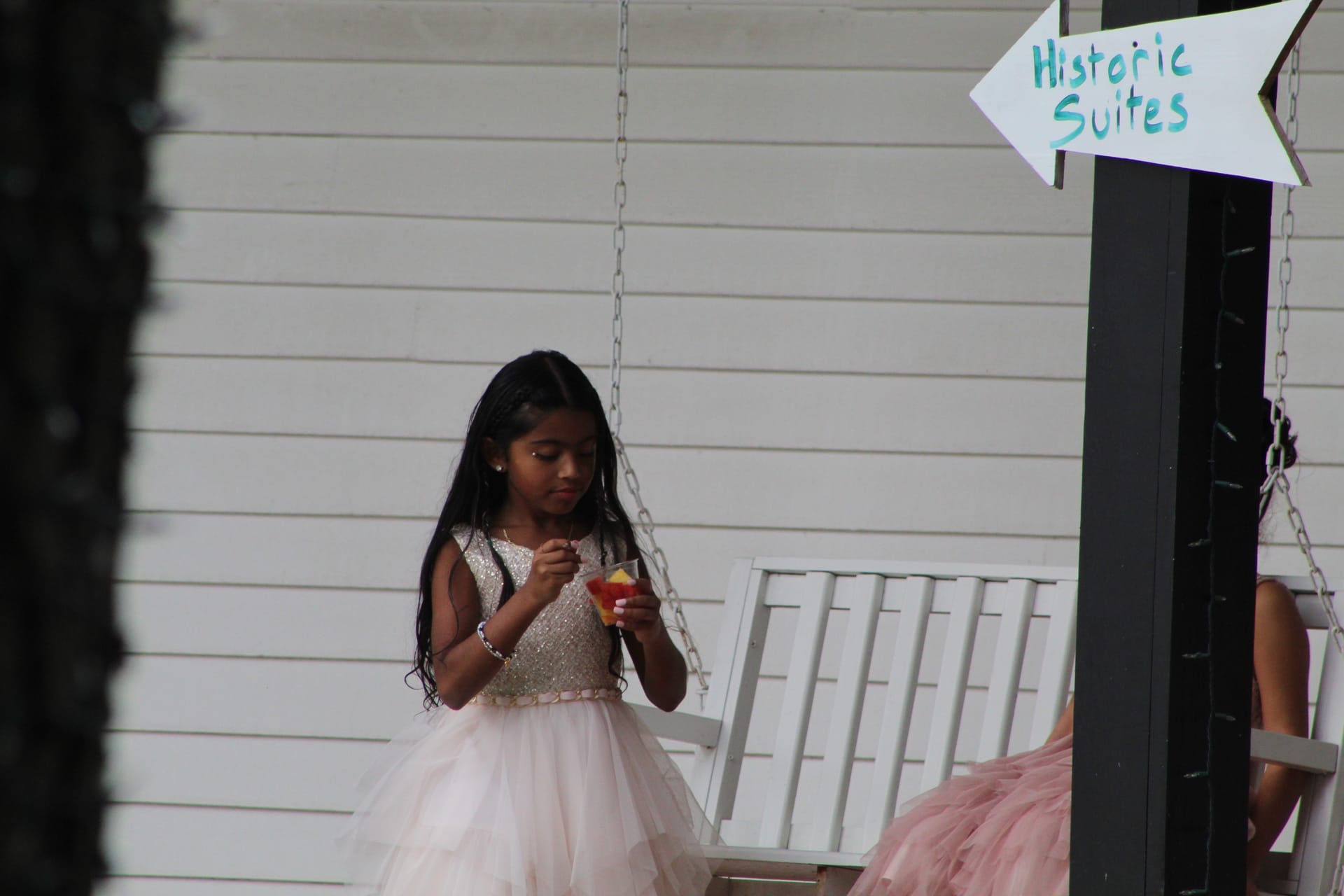 Young flower girl in a blush and gold tulle dress holding a fruity drink near a Historic Suites sign on the white porch swing at Scarborough House.