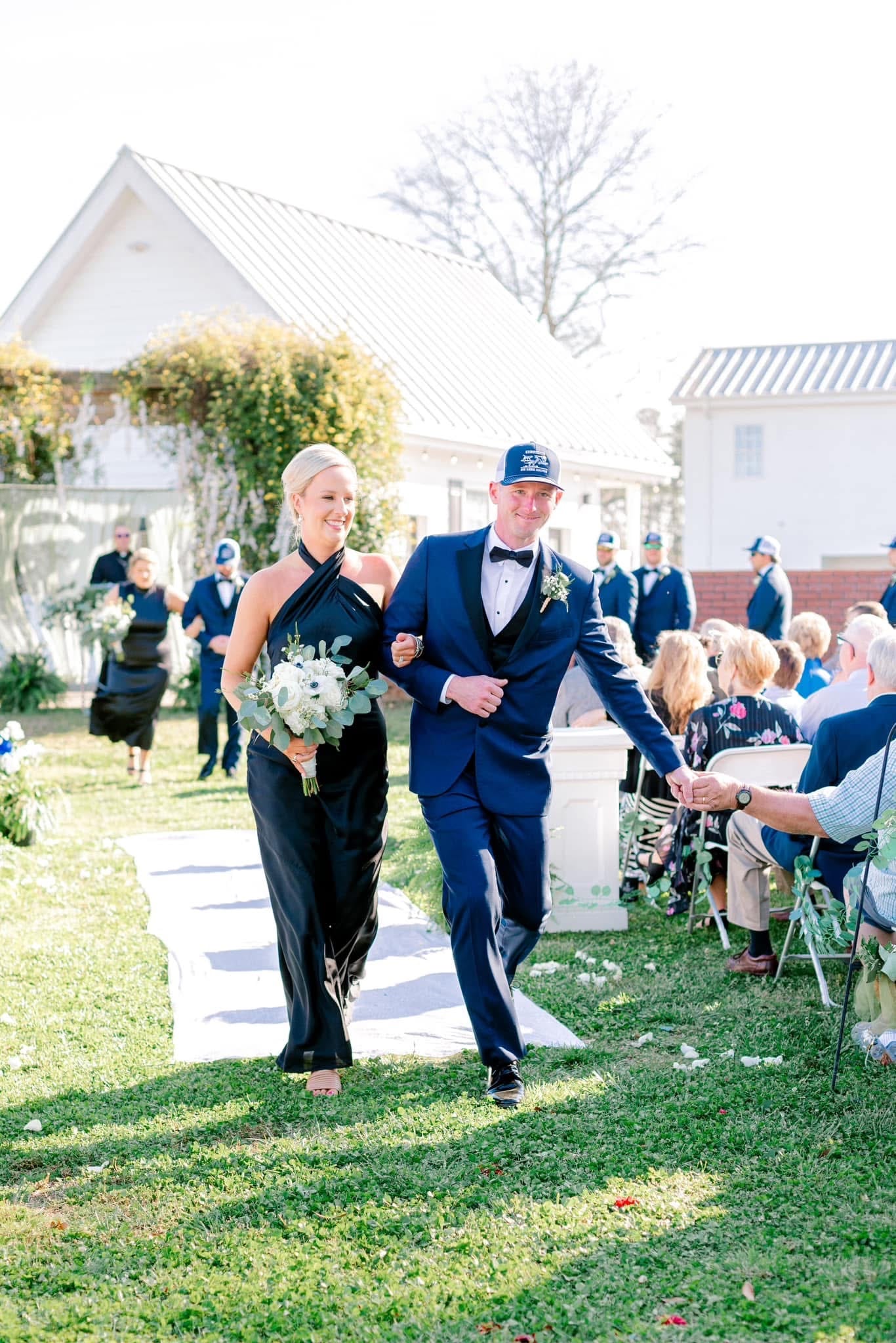 A smiling bridesmaid in a black halter gown carrying a white floral bouquet walks arm-in-arm with a groomsman in a navy tuxedo down the outdoor ceremony aisle at Scarborough House historic estate.