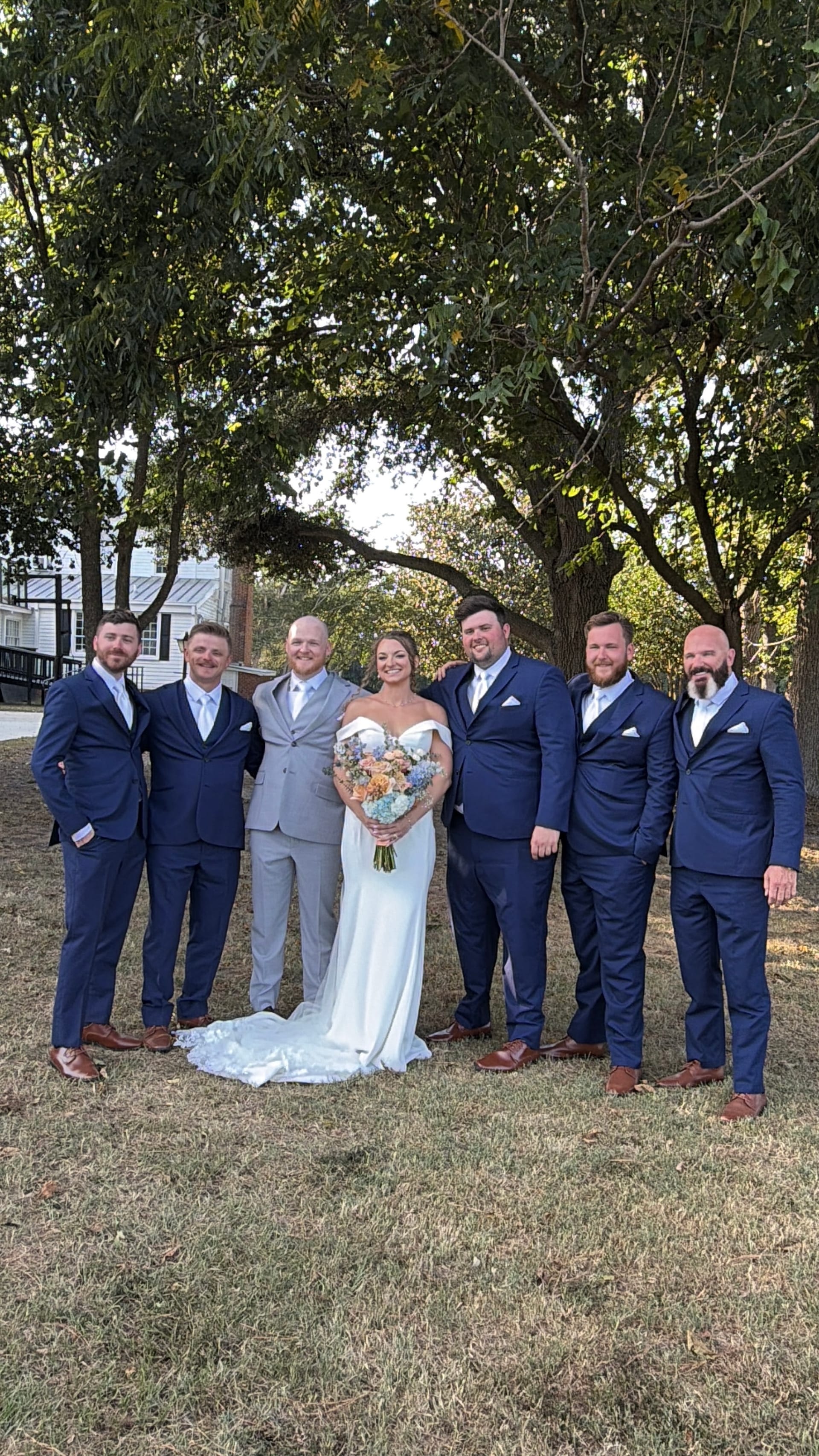 Bride in off-shoulder white gown holding a pastel bouquet stands with five groomsmen in navy suits beneath sweeping oak trees on the ceremony lawn at Scarborough House.