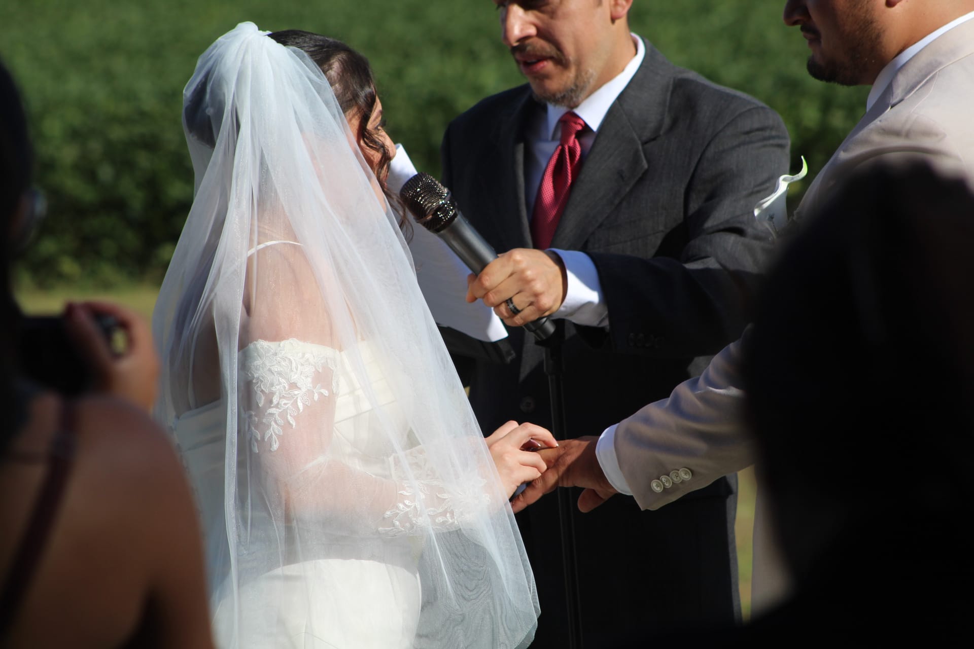 A bride in a lace wedding gown and white veil exchanges rings with the groom while an officiant holding a microphone presides over an outdoor wedding ceremony.
