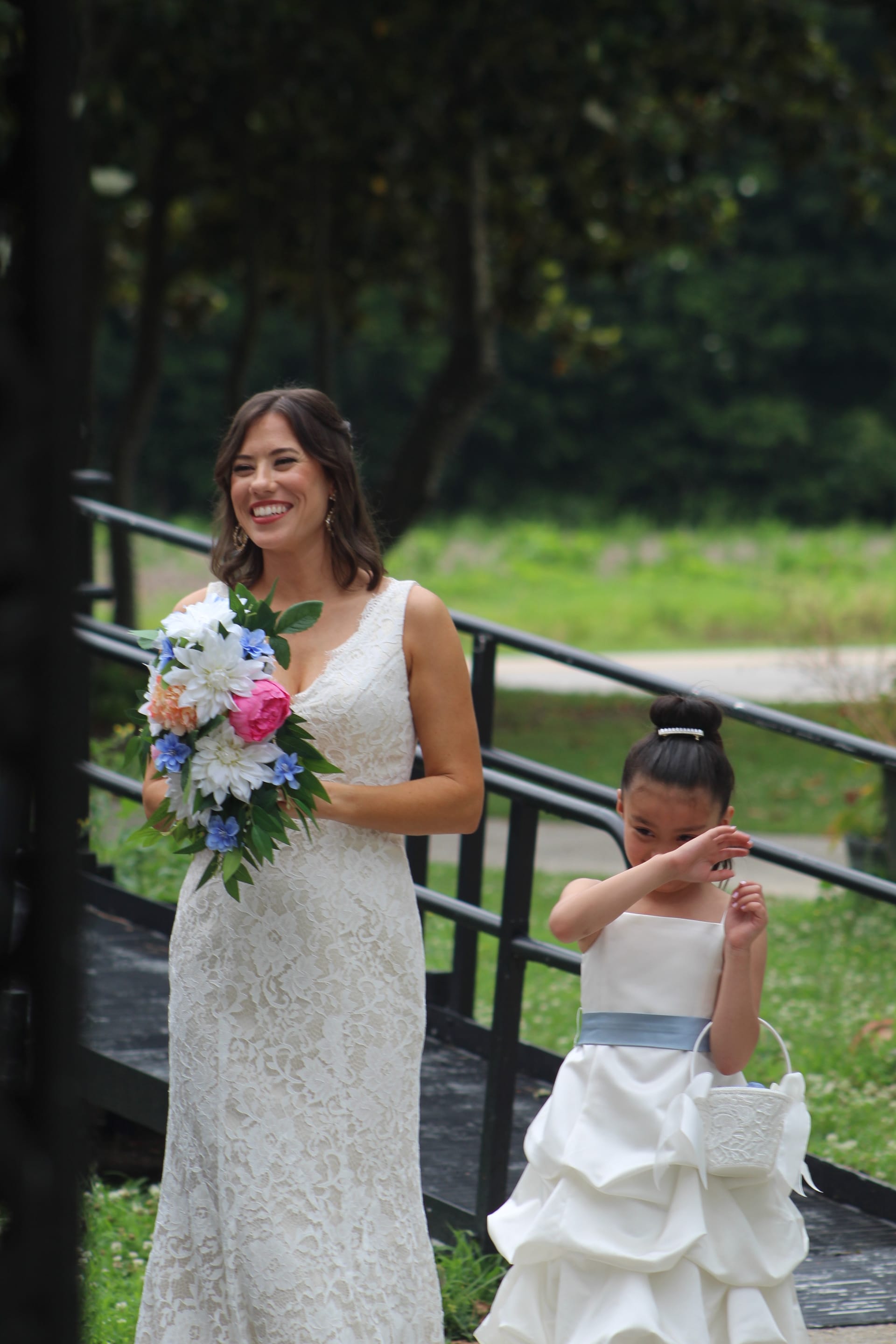 A smiling bride in a lace gown holding a colorful bouquet walks alongside a young flower girl in a white dress on a wooden bridge surrounded by lush greenery at Scarborough House.