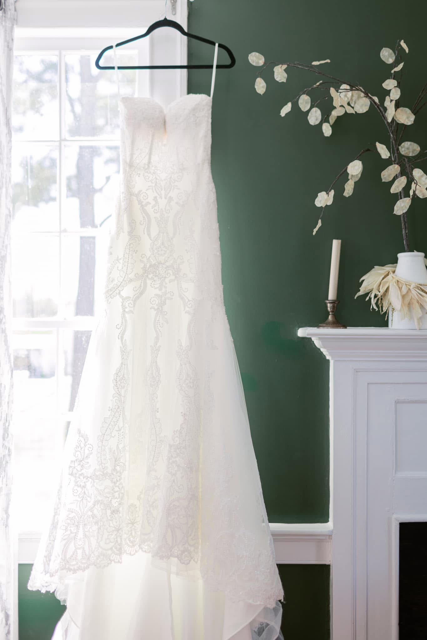 An ivory lace wedding gown hanging on a velvet hanger in front of a bright window beside a hunter-green wall and white fireplace mantle adorned with a candle and dried botanical arrangement at Scarborough House.
