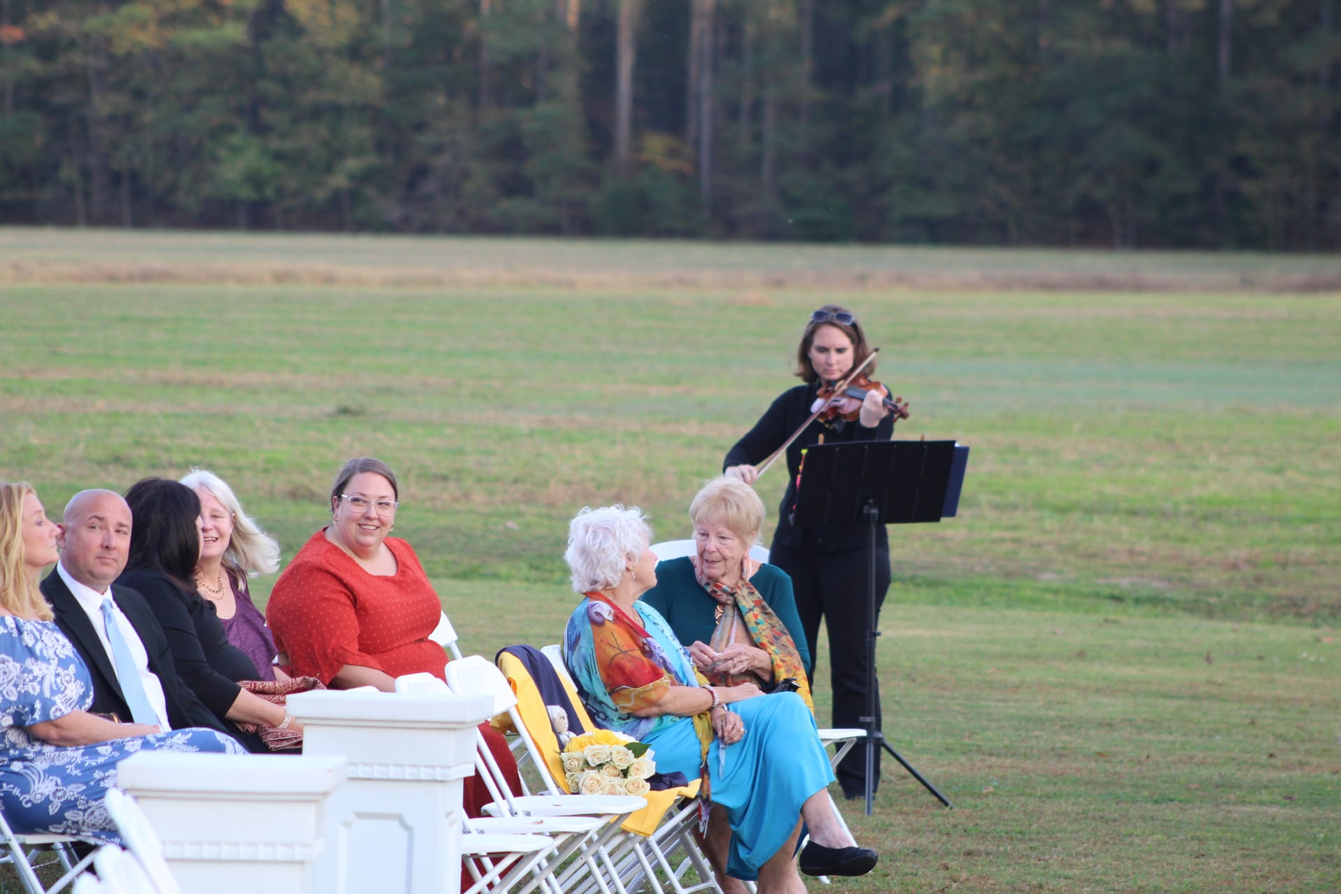 Wedding guests seated in white chairs on the open ceremony lawn listen to a violinist performing during an outdoor wedding ceremony at Scarborough House historic estate.