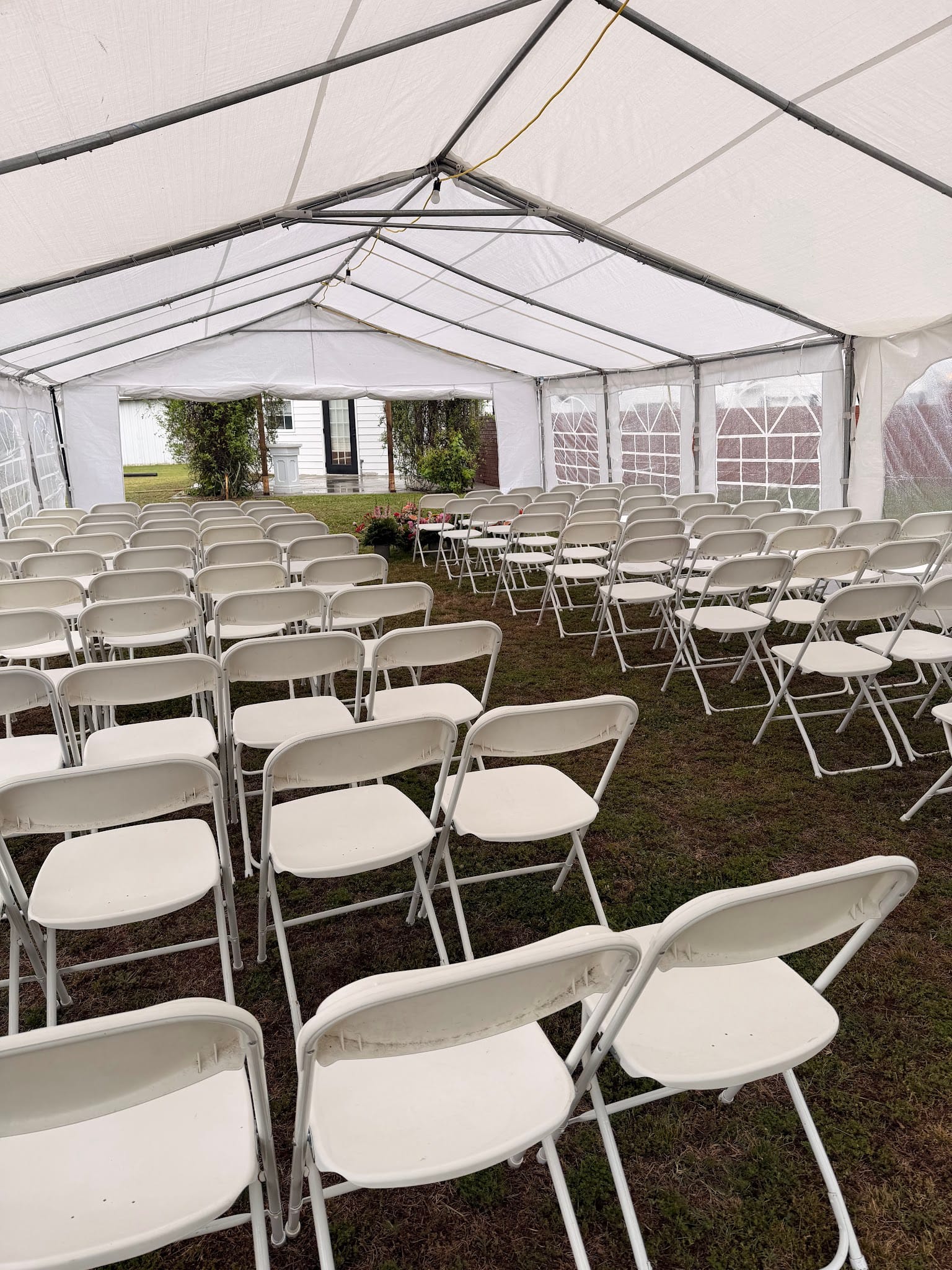 Rows of white folding chairs arranged under a large white frame tent on a grass lawn at Scarborough House estate wedding venue, with views of the historic manor house through the clear tent walls.