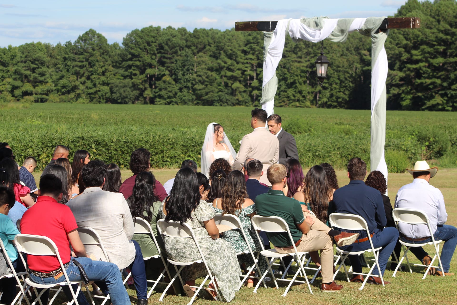 Wedding ceremony on the ceremony lawn at Scarborough House with guests seated in white folding chairs facing a couple exchanging vows beneath a rustic wooden arch draped in white fabric, surrounded by green farmland and trees.