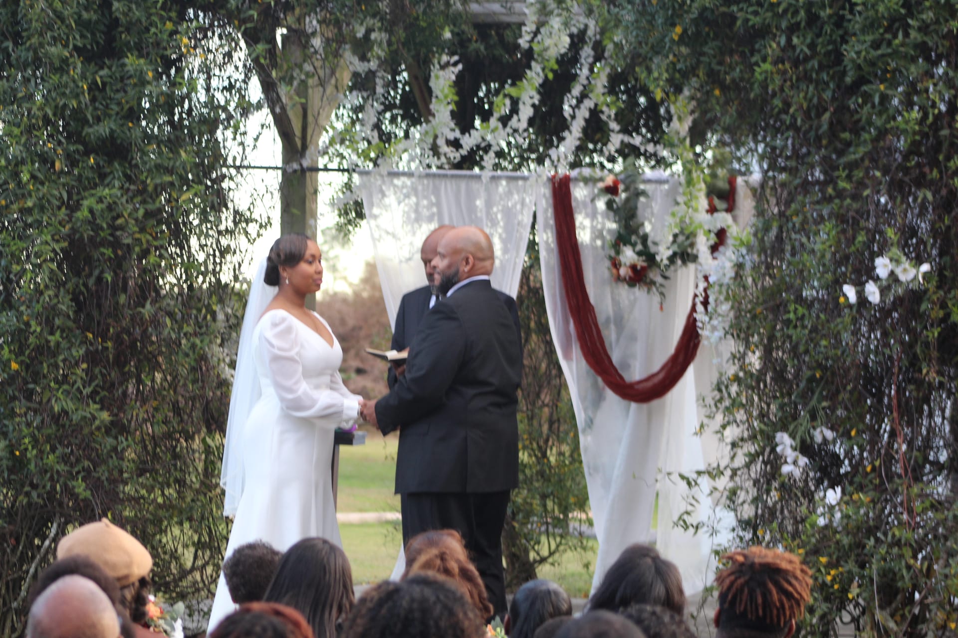 Bride in white gown and veil holds hands with groom in black suit beneath a decorated arch draped in white fabric and burgundy sash during an outdoor garden wedding ceremony at Scarborough House.