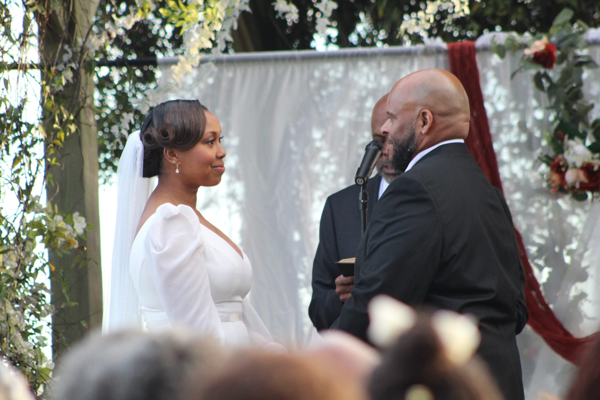Bride in a white gown and veil gazes at her groom during their outdoor wedding ceremony beneath a floral-draped arch at Scarborough House.