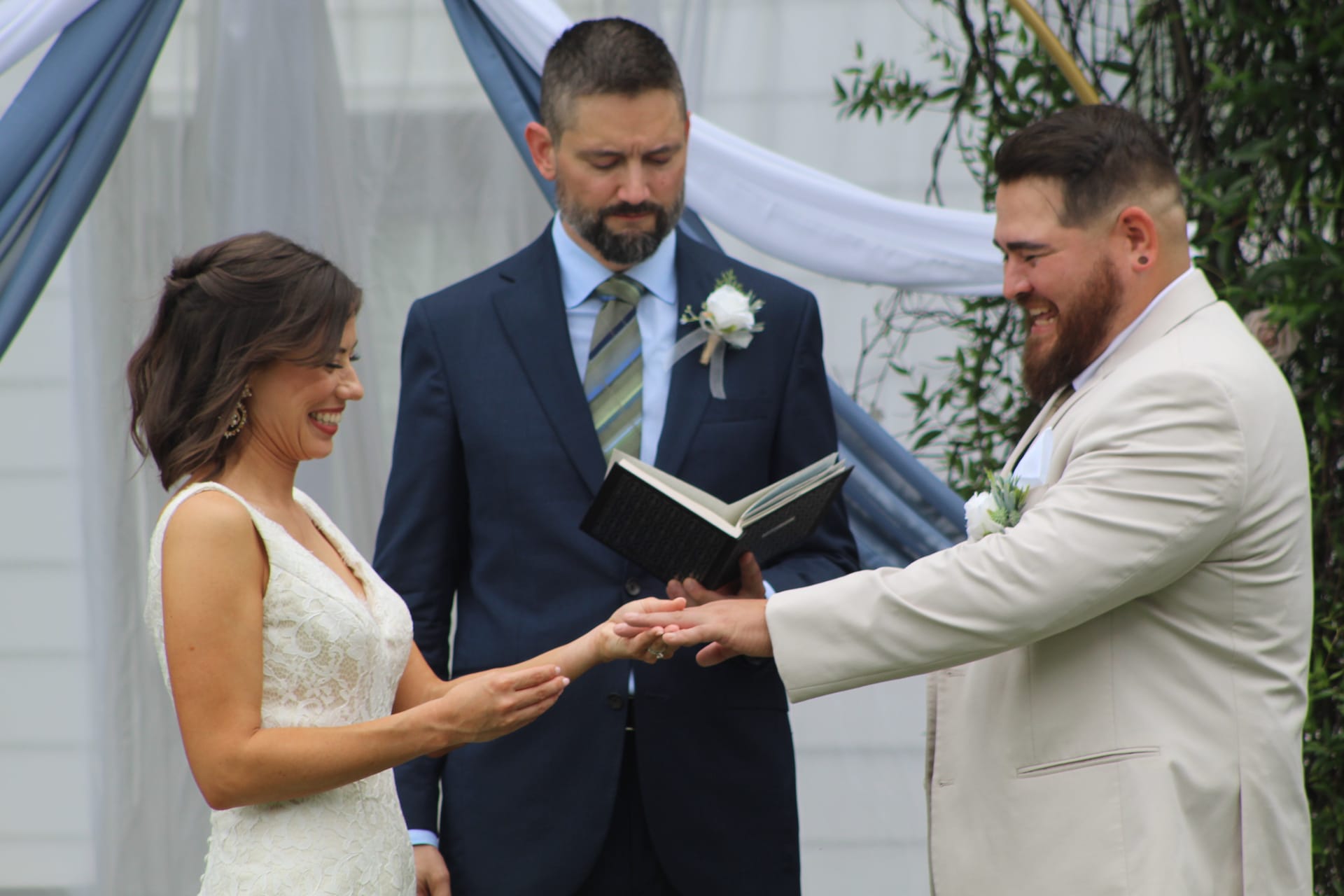 Smiling groom places wedding ring on brides finger during outdoor ceremony beneath a blue and white draped arch while the officiant reads from a book.