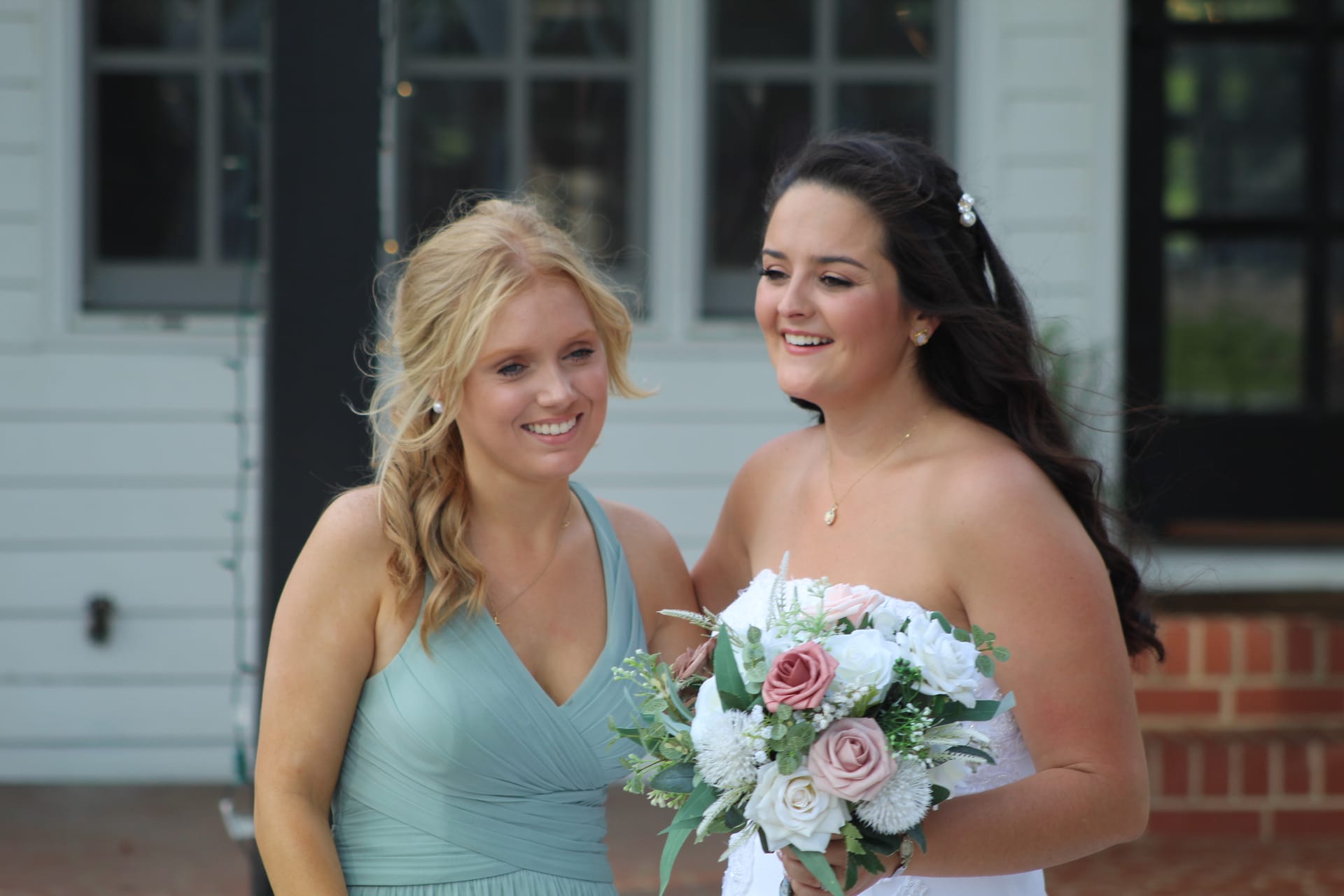Bride in a strapless white gown holding a pink and white floral bouquet smiles with her bridesmaid in a sage green dress outside the historic Scarborough House manor.