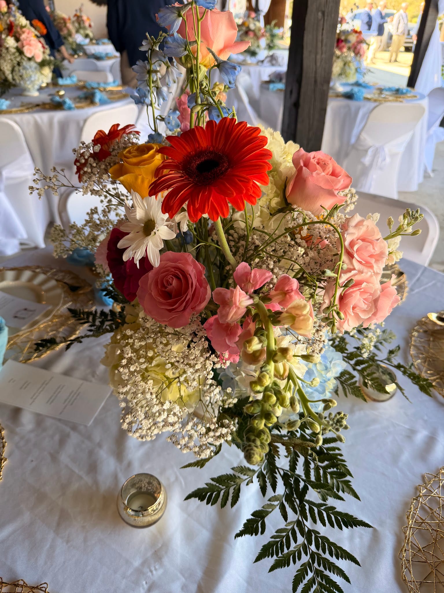 A lush wedding reception centerpiece featuring red gerbera daisies, pink roses, baby's breath, and blue delphinium on a white linen table at Scarborough House.