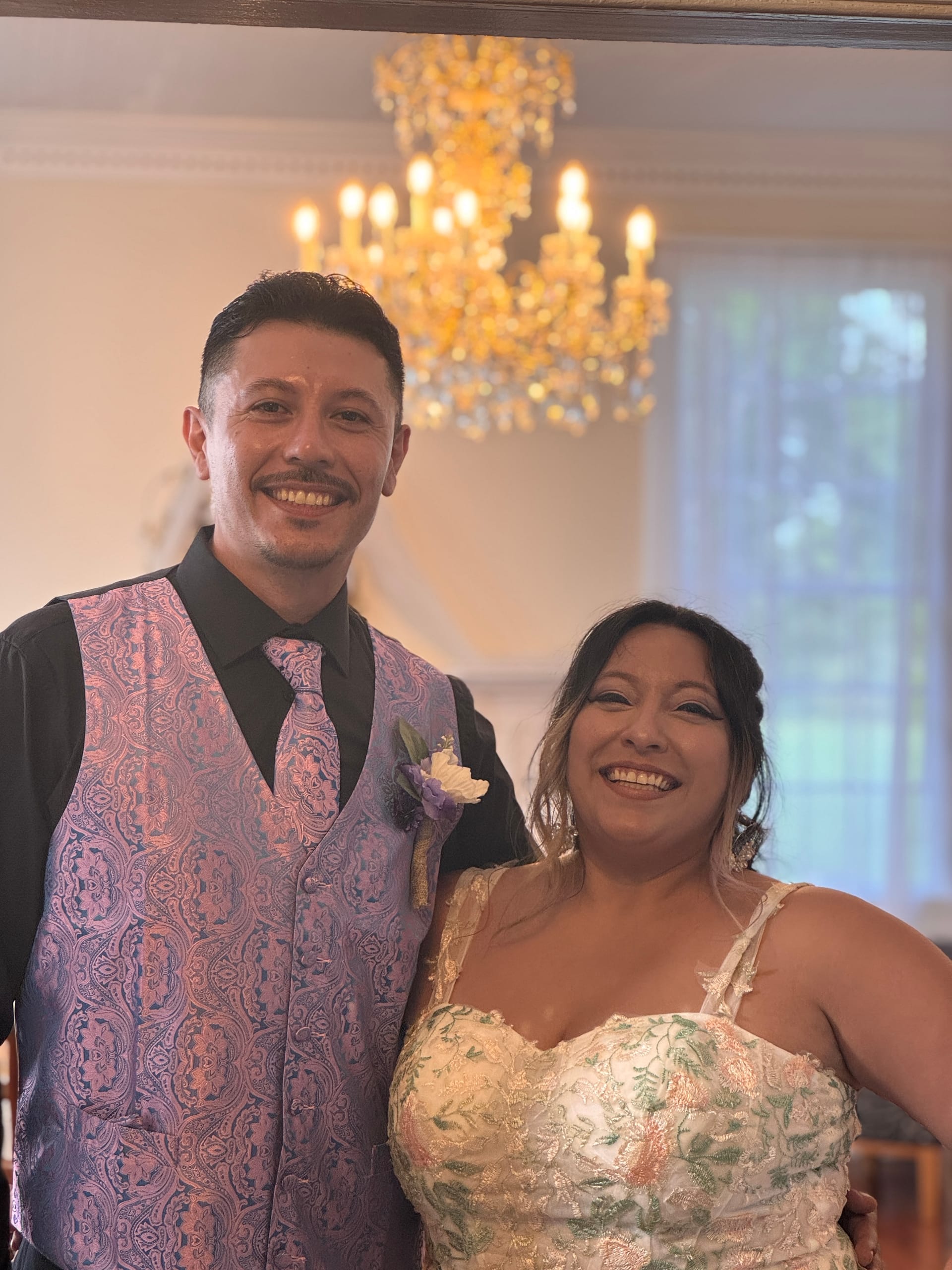 Smiling bride and groom posing together beneath a golden chandelier in the grand hall of Scarborough House, he wearing a pink paisley vest with a boutonniere and she in a floral embroidered gown.