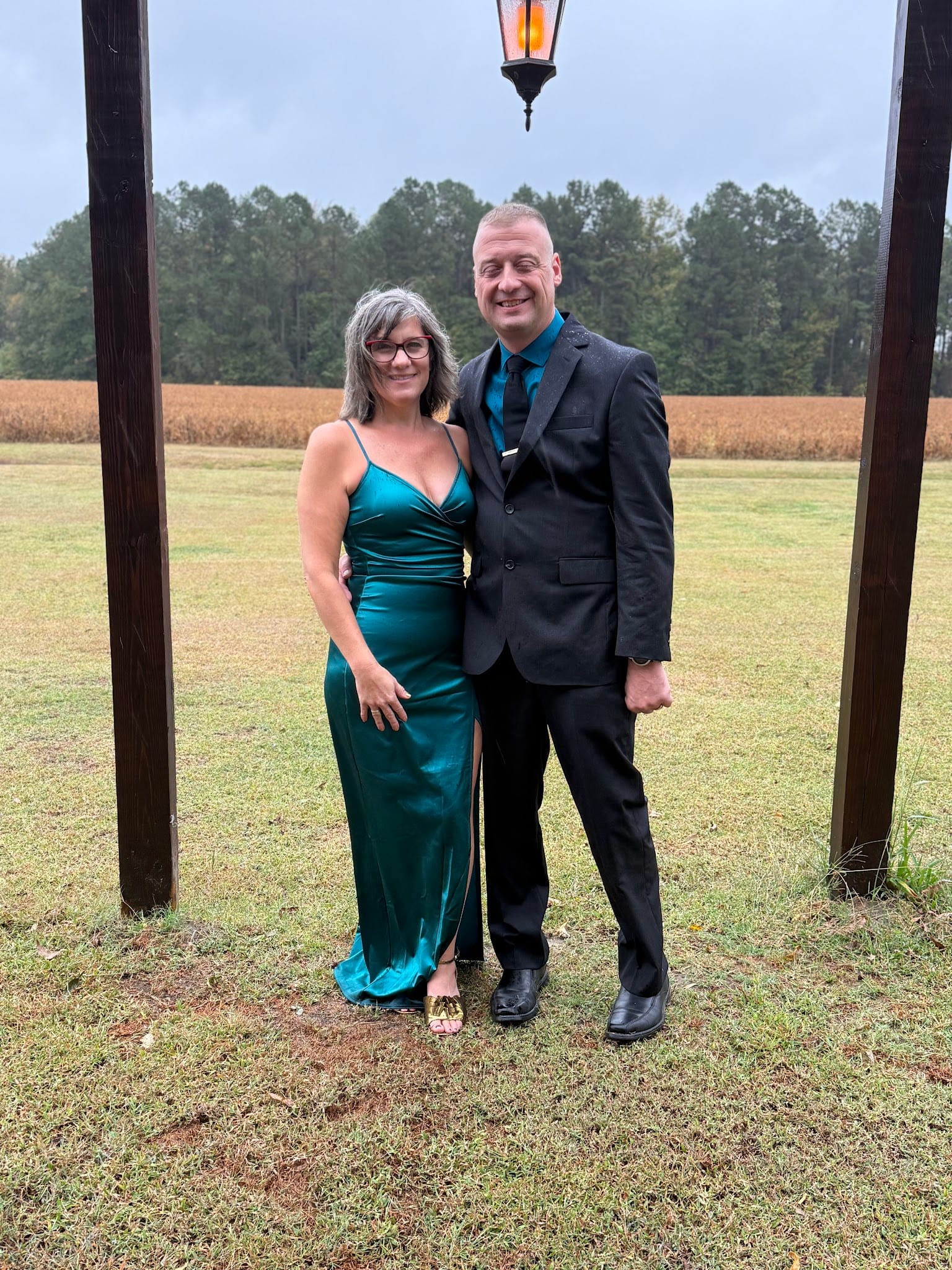 Elegantly dressed couple posing under a hanging lantern with Scarborough House farmland fields and pine tree line visible in the background on a cloudy day.