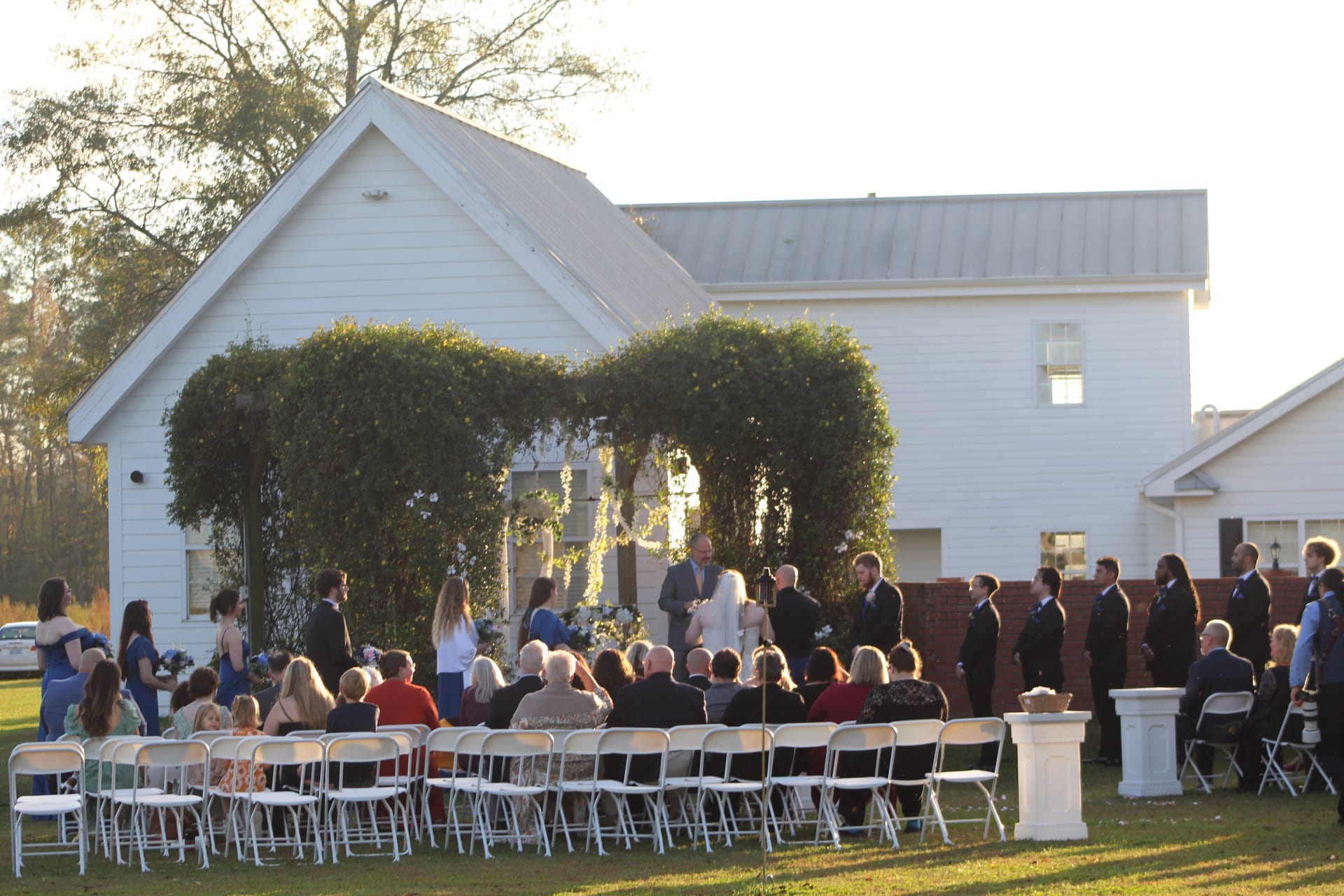Outdoor wedding ceremony at golden hour on the ceremony lawn of Scarborough House with guests seated in white folding chairs facing the couple at an ivy-draped white historic farmhouse altar.