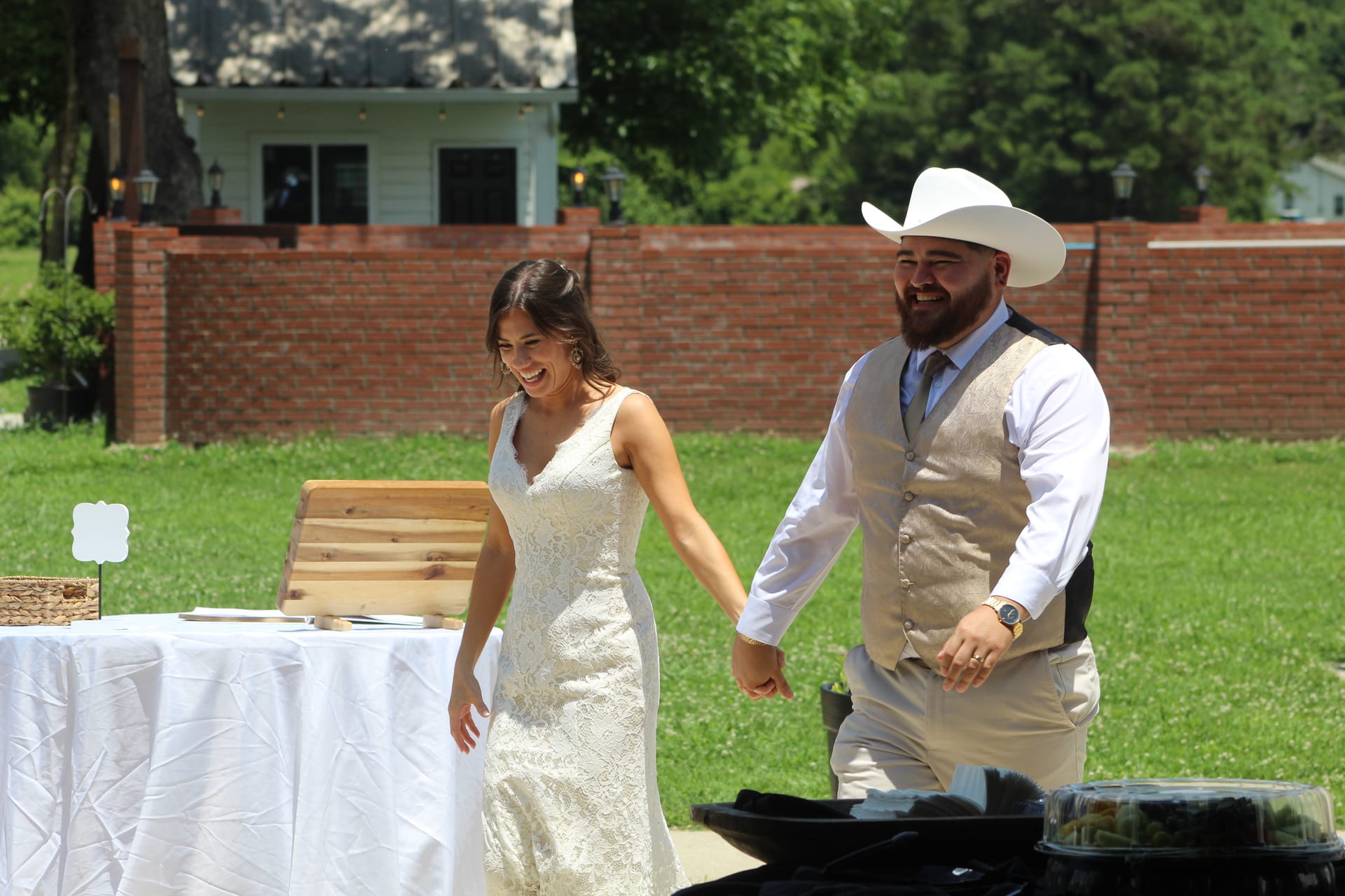 Smiling bride in a lace gown and groom in a tan vest and white cowboy hat walk hand-in-hand across the sunlit lawn at Scarborough House wedding venue.