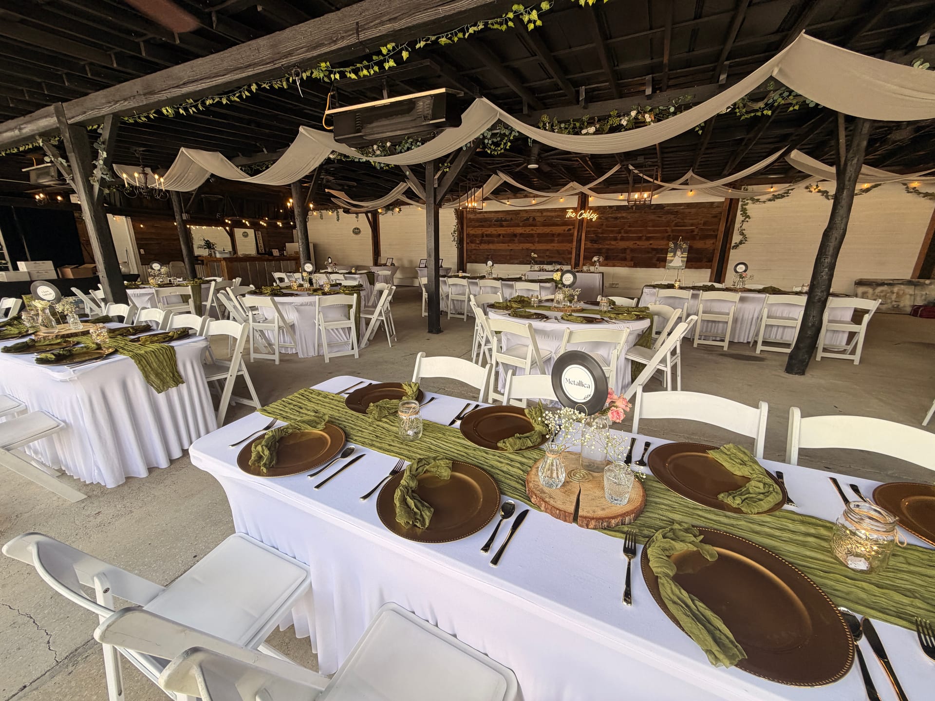 Elegantly styled wedding reception tables under a draped open-air pavilion featuring bronze charger plates, sage green napkins, wood-slice centerpieces, and string lights at Scarborough House.