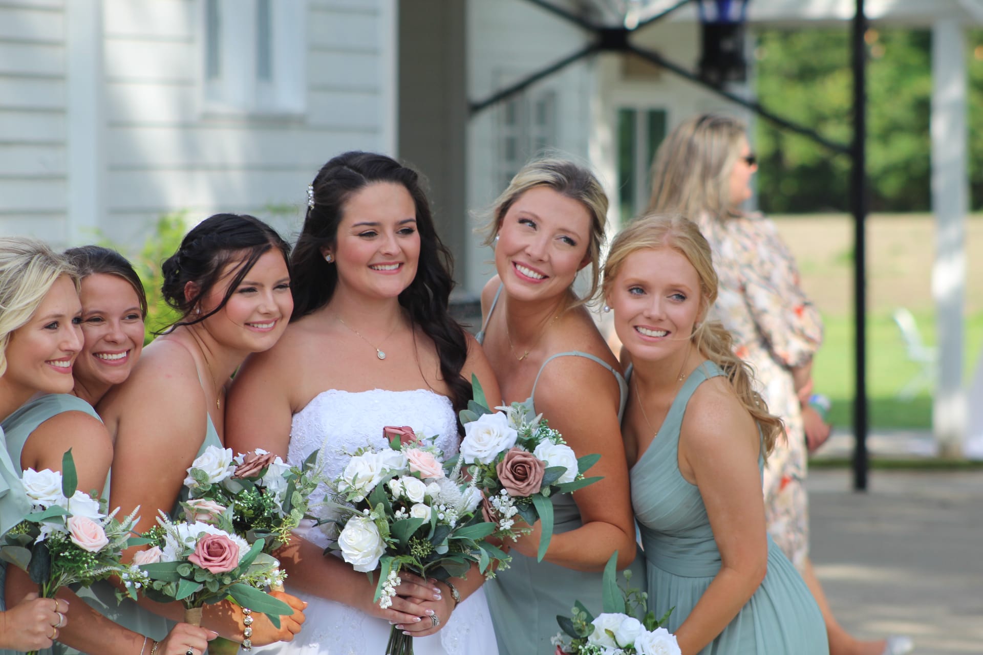 Bride in white strapless gown poses and laughs with five bridesmaids in sage green dresses holding white and blush bouquets in front of the historic Scarborough House manor.