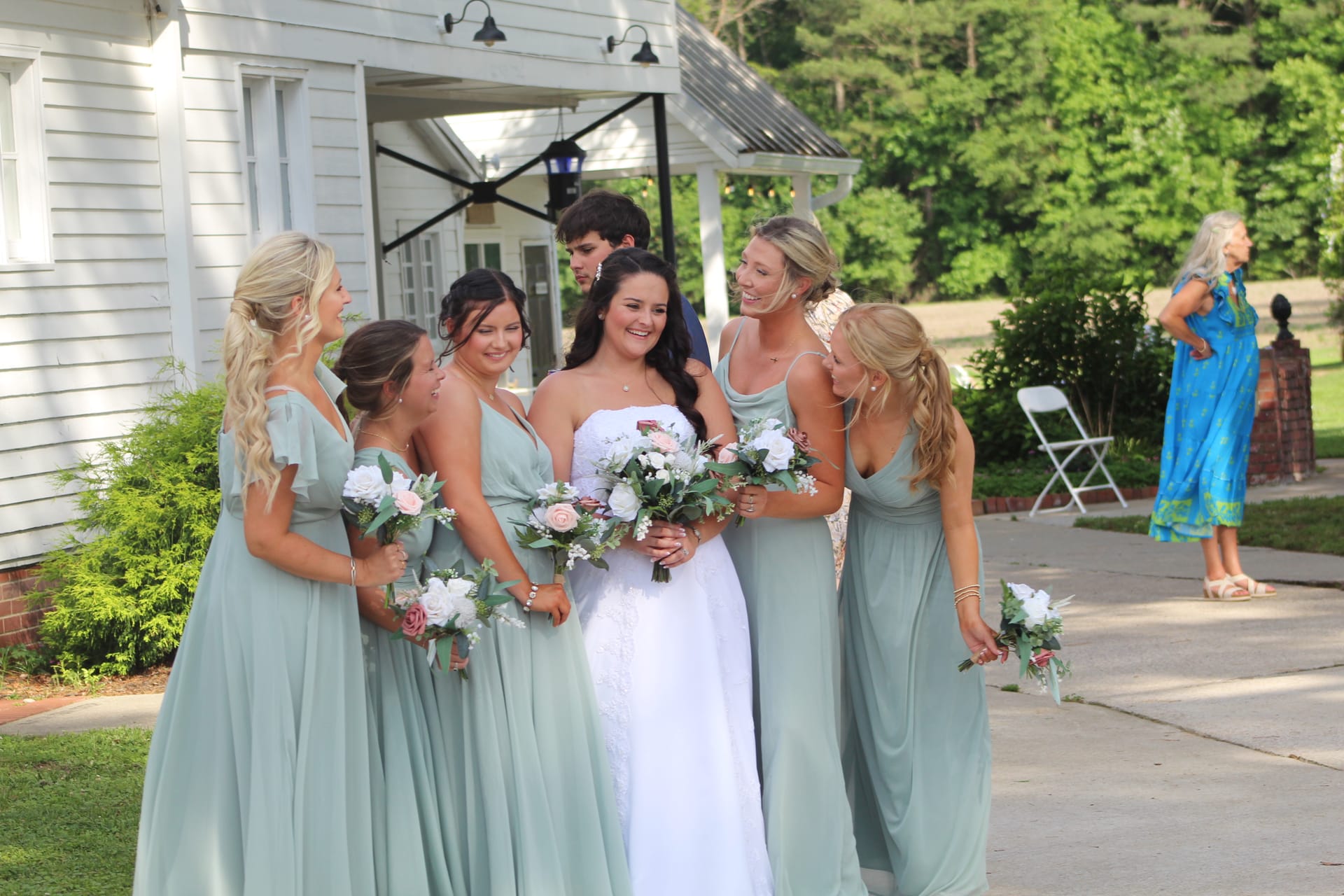 Bride in white lace gown laughing with five bridesmaids in sage green dresses holding floral bouquets outside the historic Scarborough House manor.