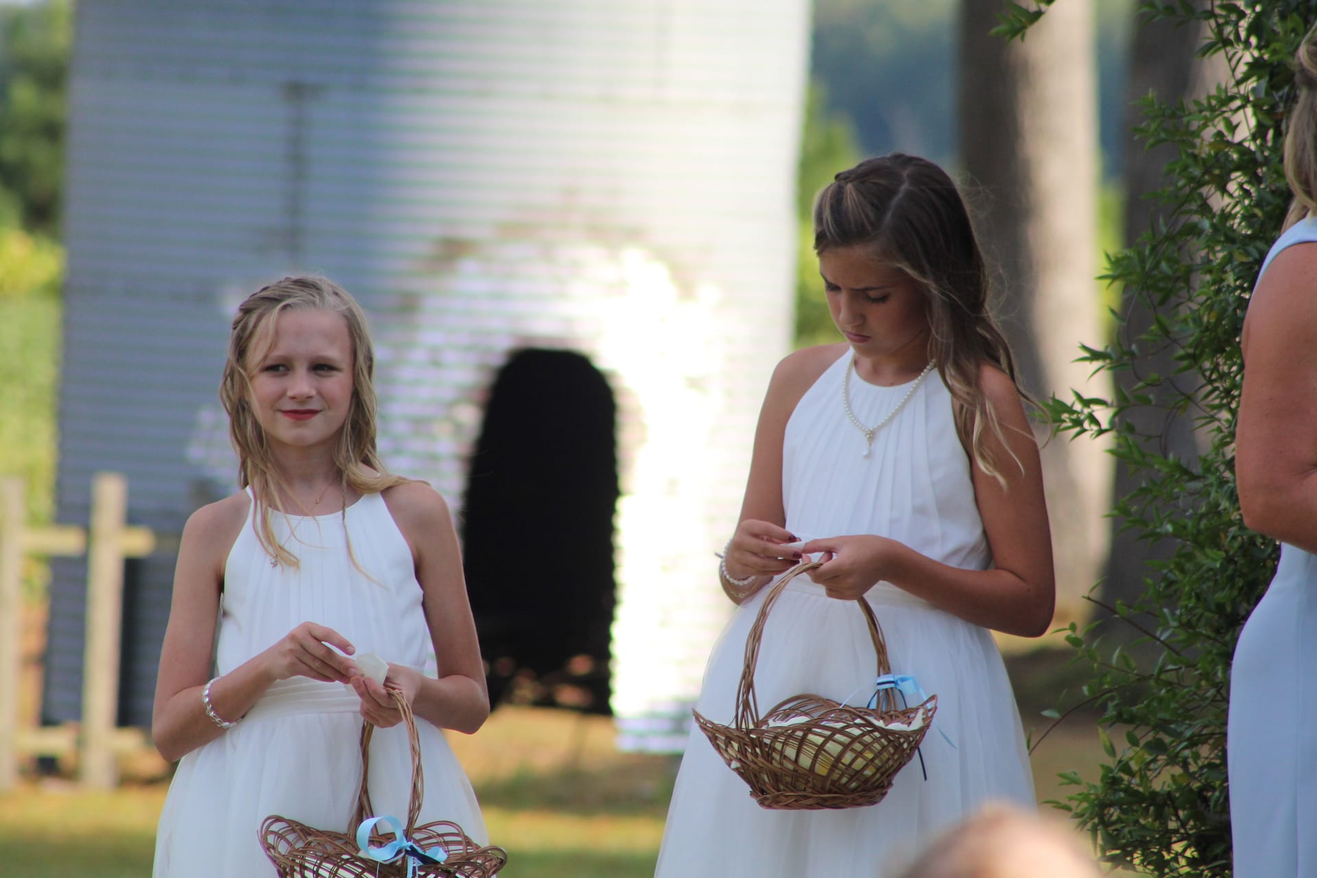 Two young flower girls in white halter dresses carrying wicker baskets walk down the outdoor ceremony aisle at Scarborough House with a historic white silo glowing in the golden background.